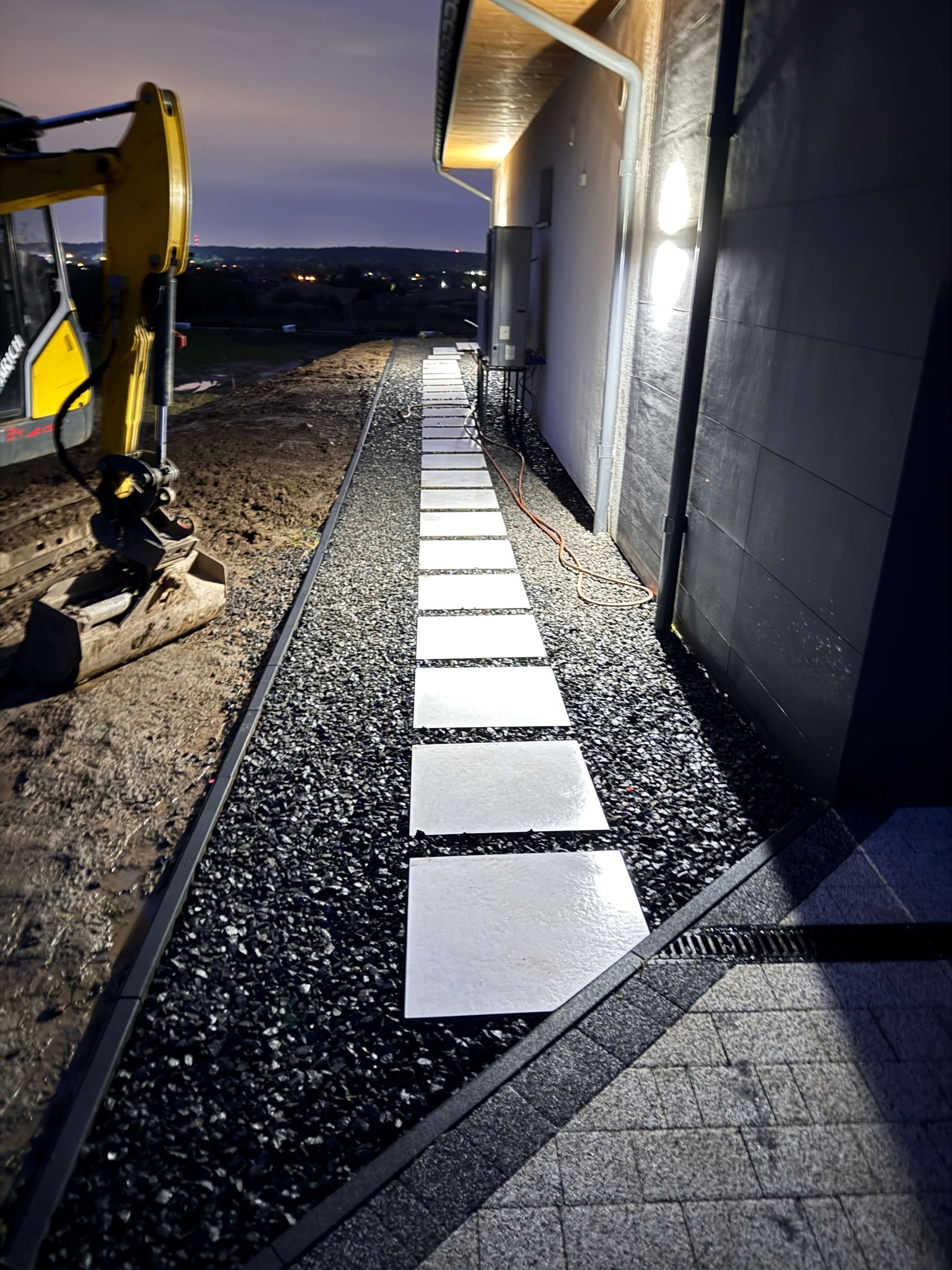 Nowoczesna ścieżka z białych płyt ułożona na czarnym żwirze, oświetlona lampami przy ścianie budynku. W tle koparka i wieczorny krajobraz.