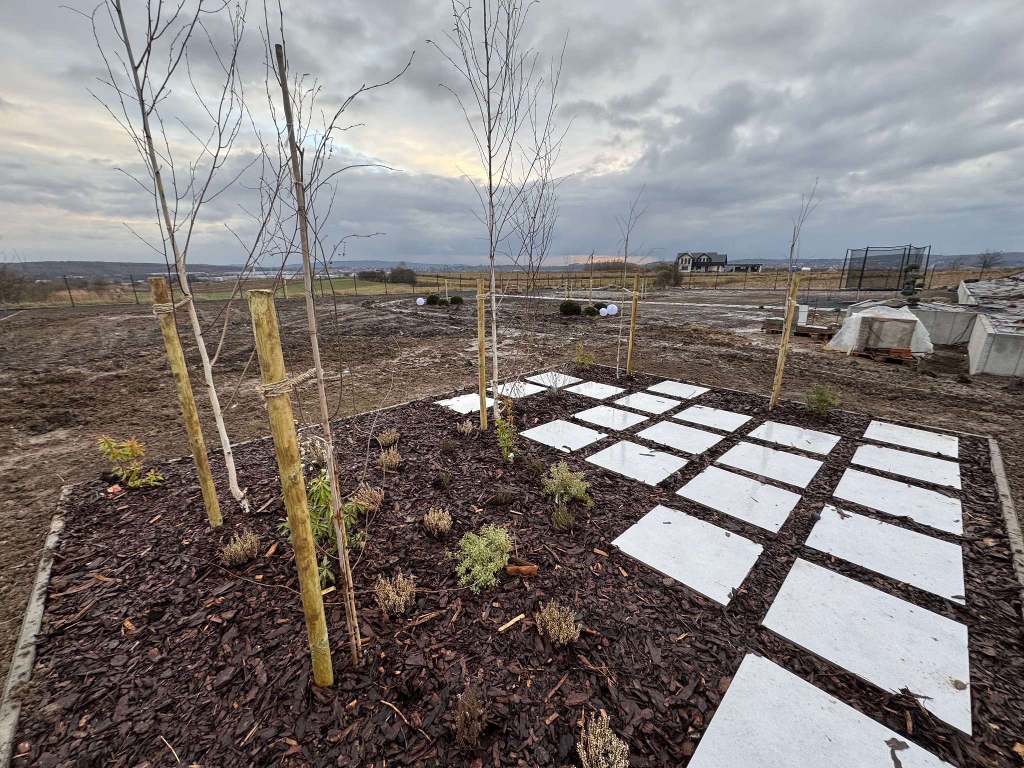 Fragment ogrodu po realizacji: młode drzewka podparte drewnianymi palikami, rabata z korą i nasadzeniami, kwadratowe płyty chodnikowe. Pochmurne niebo w tle.
