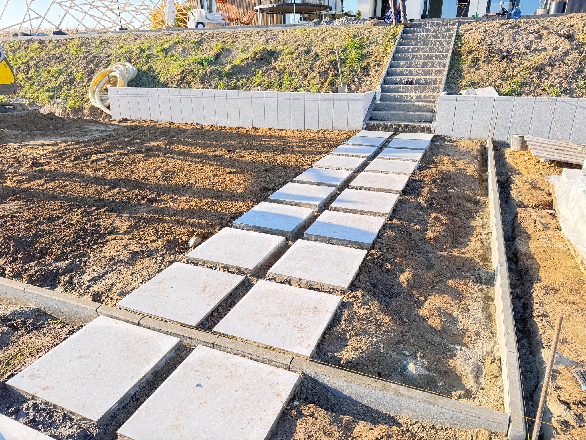 Układanie ścieżki z płyt na gruncie, widoczne obrzeża i schody w tle. Prace ziemne przygotowujące teren pod przyszły ogród.