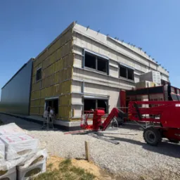 Budynek w trakcie ocieplania elewacji z widocznym rusztowaniem i materiałami izolacyjnymi. Dwóch pracowników na drabinie. Prace w toku, jasne niebo.