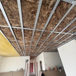 MS.Maryna Shcherbatiuk - Poddasze z izolacją z wełny mineralnej i metalową konstrukcją sufitu podwieszanego, częściowo zakryte folią paroizolacyjną. Widok z dołu, w tle pomieszczenie w trakcie remontu.