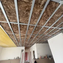 Poddasze z izolacją z wełny mineralnej i metalową konstrukcją sufitu podwieszanego, częściowo zakryte folią paroizolacyjną. Widok z dołu, w tle pomieszczenie w trakcie remontu.