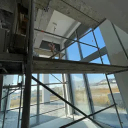 Malowanie sufitu w jasnym wnętrzu na rusztowaniu, widoczne duże okna z widokiem na zewnątrz. Praca w toku, sufit częściowo pomalowany, widoczne ślady farby.