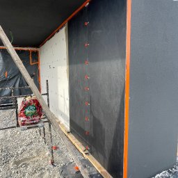 MS.Maryna Shcherbatiuk - Fragment elewacji w trakcie prac: biała izolacja, czarna membrana, pomarańczowe profile. Widoczne rusztowanie i gruz. Etap przed finalnym wykończeniem.