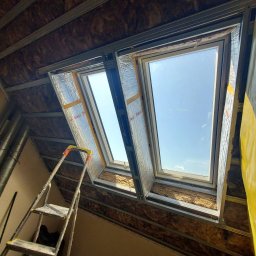 MS.Maryna Shcherbatiuk - Dwa okna dachowe w trakcie montażu, widoczne ocieplenie wełną mineralną i metalowa konstrukcja. Ustawiona drabina malarska. Widok na błękitne niebo.