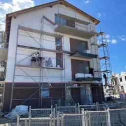 Ekipa maluje elewację dwukondygnacyjnego domu z rusztowania. Część ściany w kolorze białym, część brązowym. Błękitne niebo w tle. Widoczne narzędzia i materiały budowlane.