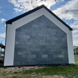 Elewacja domu z szarej, kamiennej okładziny, białe obramowanie, rusztowanie z boku. Słońce i chmury na niebie. Widok z dołu, podkreślający geometrię budynku.