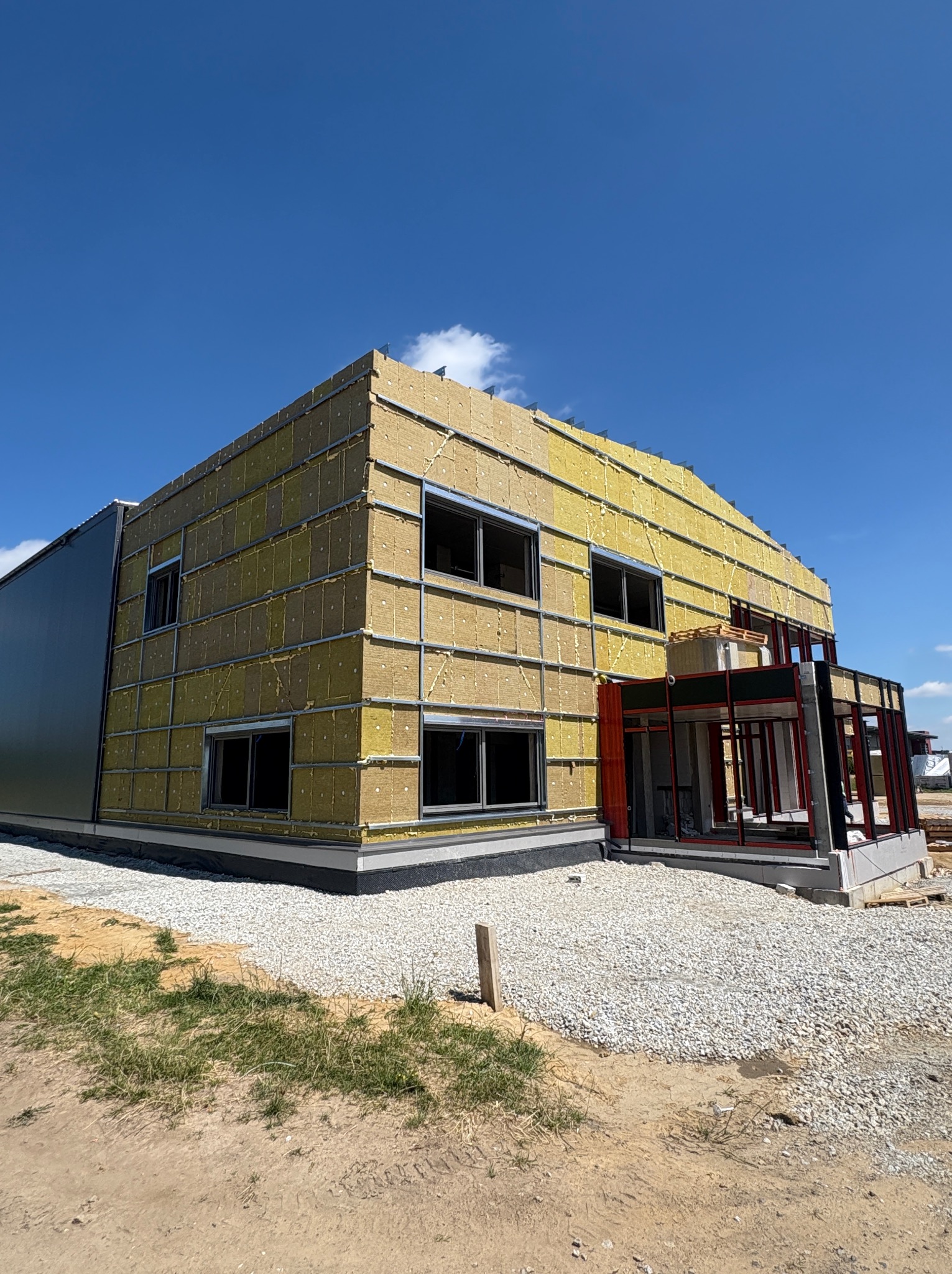 Budynek w trakcie ocieplania z widoczną warstwą izolacji i metalową konstrukcją. Widoczne okna i częściowo ukończona elewacja. Jasne niebo w tle.