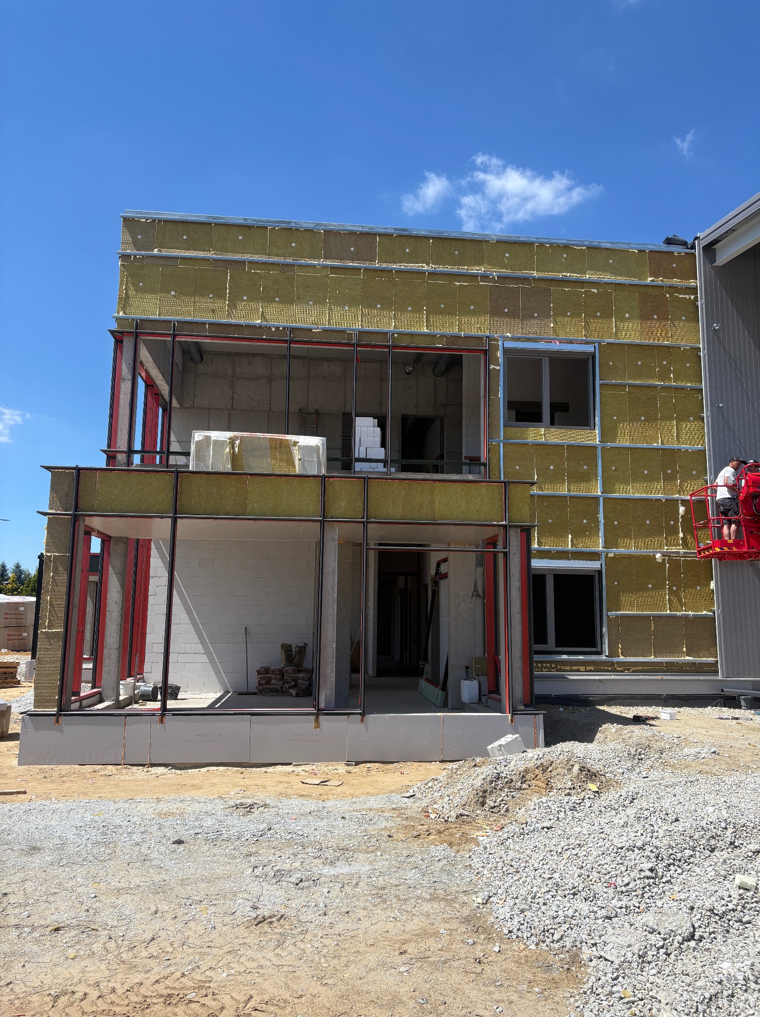 Budowa dwukondygnacyjnego domu z ociepleniem ścian zewnętrznych wełną mineralną. Widoczna konstrukcja stalowa i pracownik na podnośniku koszowym.