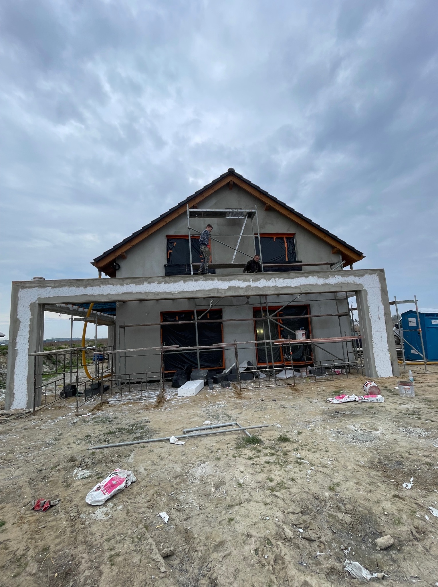 Dom w trakcie prac elewacyjnych: szare ściany, rusztowania, widoczny pracownik. Częściowo ocieplony fragment z białym styropianem. Pochmurne niebo w tle.