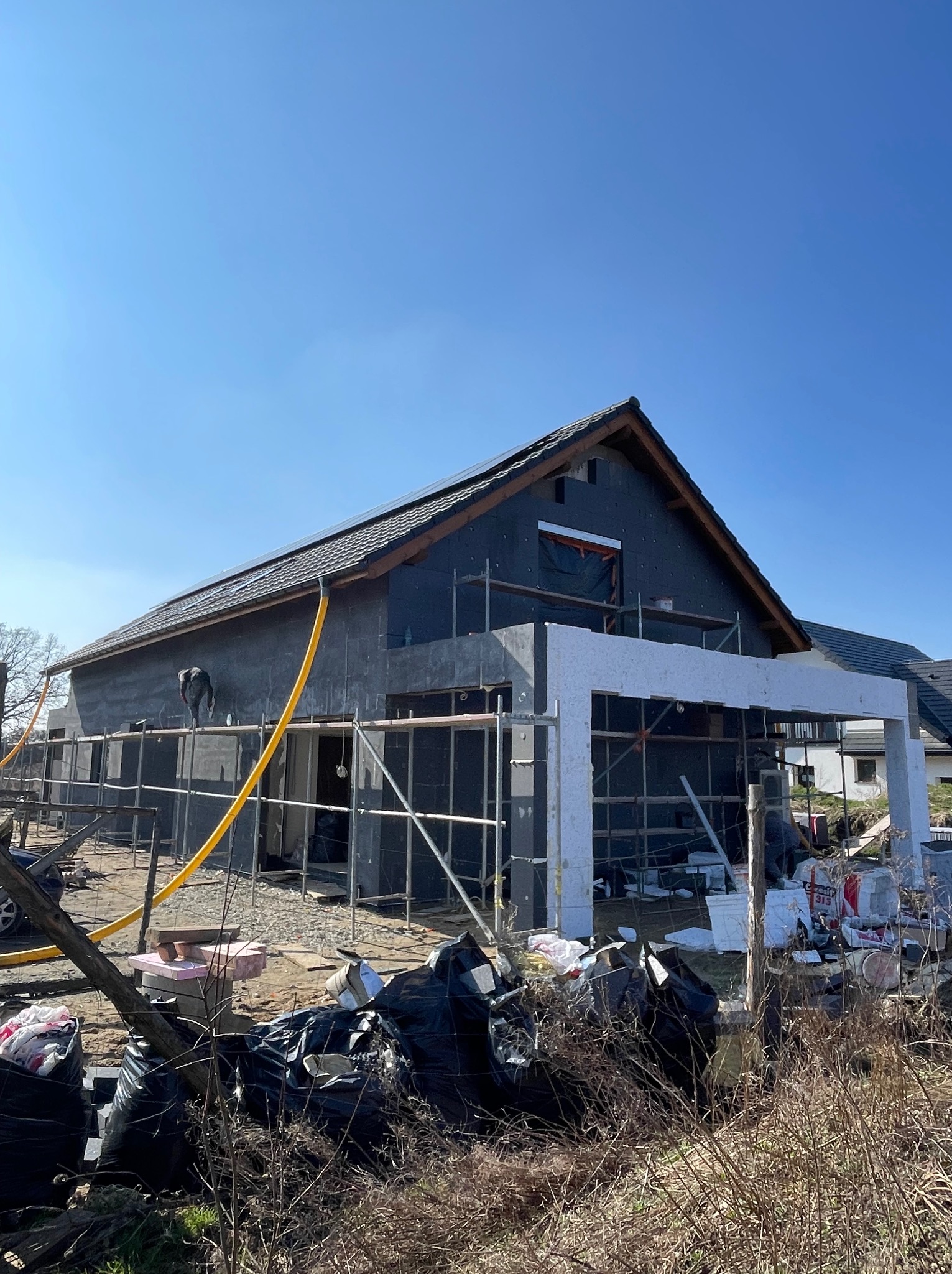 Dom w trakcie ocieplania elewacji, widoczne rusztowania, styropian i pracownik na rusztowaniu. Żółty wąż na elewacji. W tle błękitne niebo.