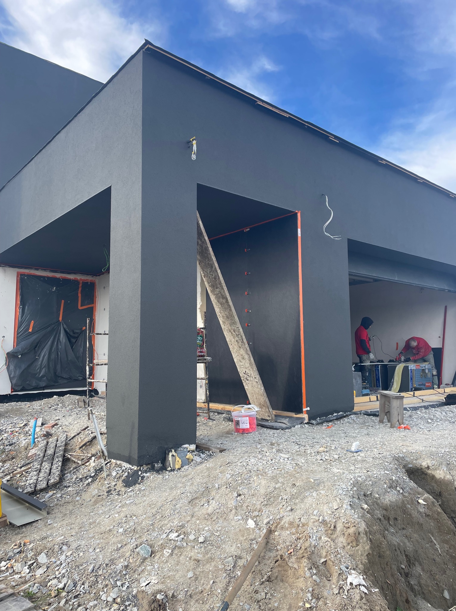 Nowoczesny dom z ciemną elewacją w trakcie prac wykończeniowych. Widoczni pracownicy, rusztowania i materiały budowlane. Ujęcie z dołu, błękitne niebo w tle.