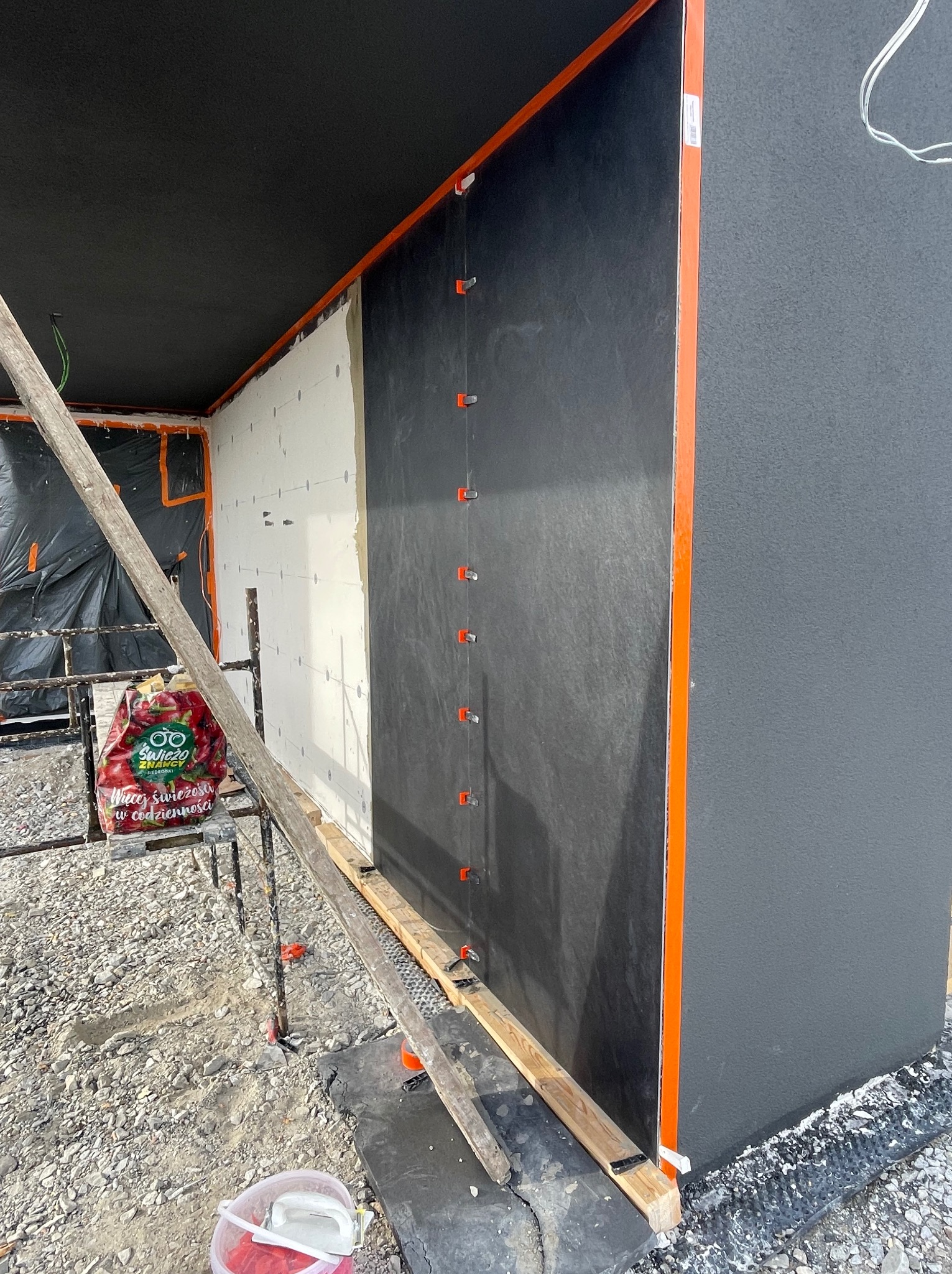 Fragment elewacji w trakcie prac: biała izolacja, czarna membrana, pomarańczowe profile. Widoczne rusztowanie i gruz. Etap przed finalnym wykończeniem.