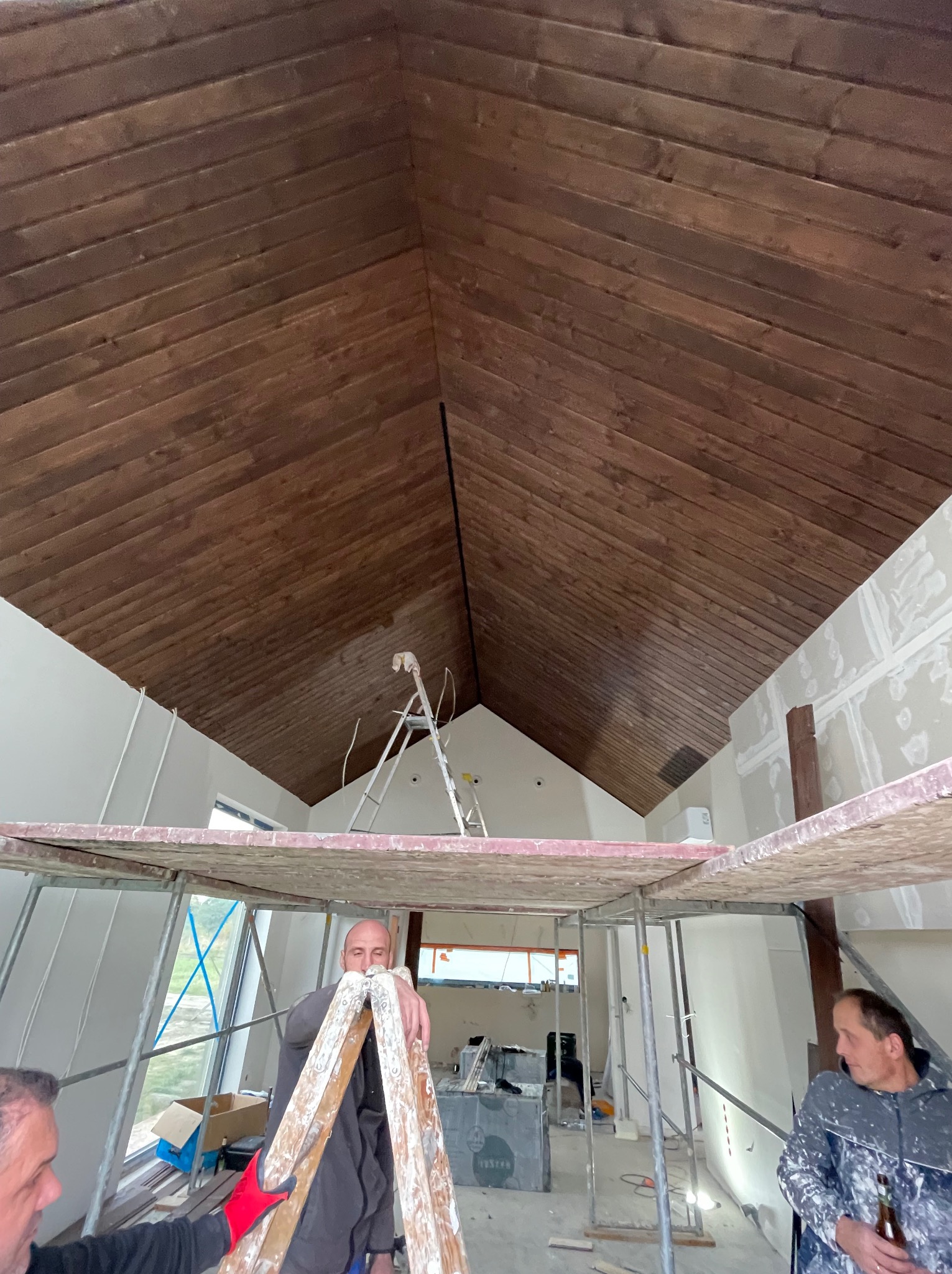 Wnętrze w trakcie prac wykończeniowych: drewniany sufit, rusztowanie, ściany przygotowane do malowania. Widoczni pracownicy w trakcie pracy.