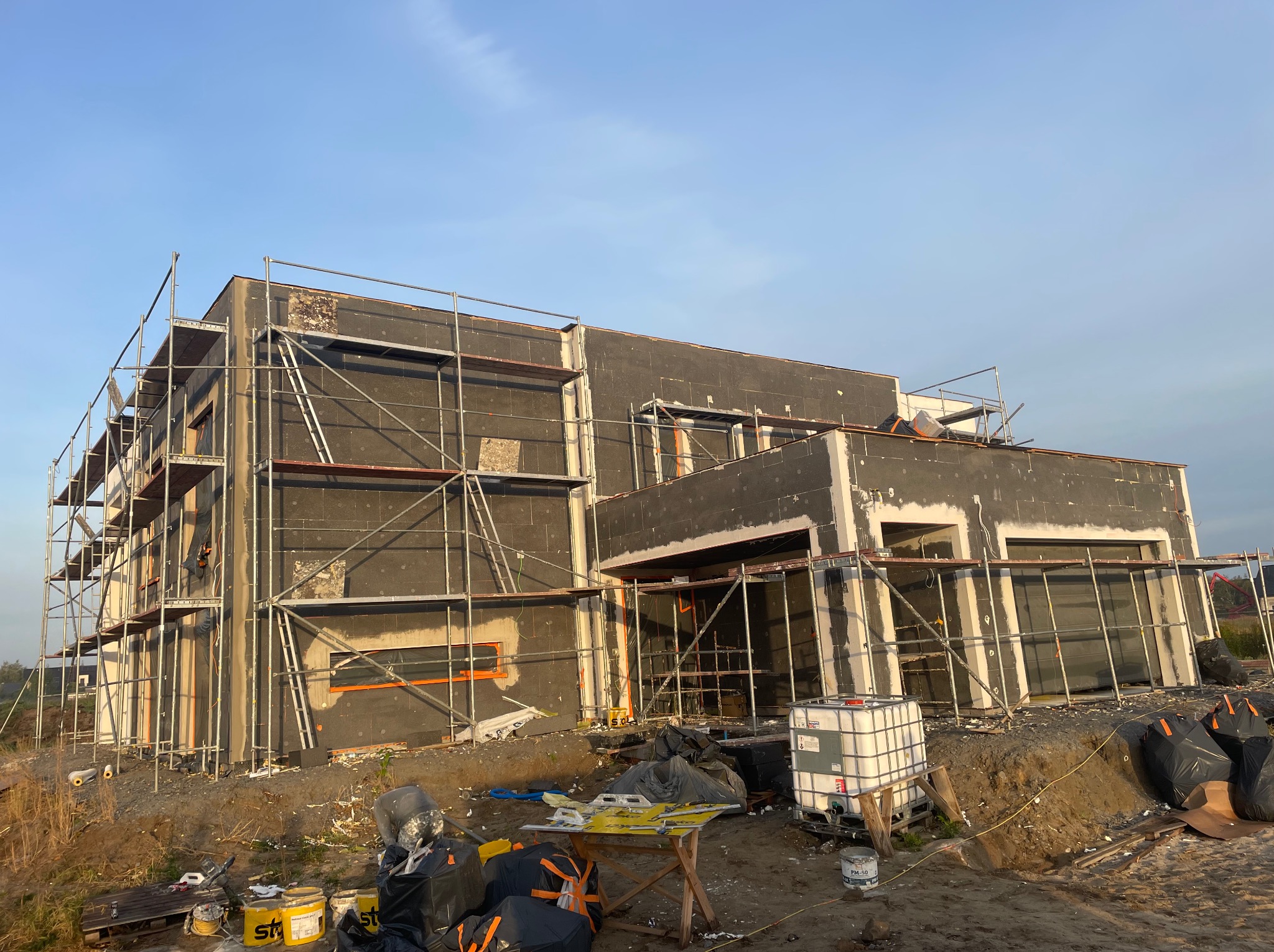 Dom w trakcie ocieplania, widoczne rusztowania i czarne płyty izolacyjne. Częściowo otynkowane fragmenty elewacji. Na pierwszym planie narzędzia i materiały budowlane.