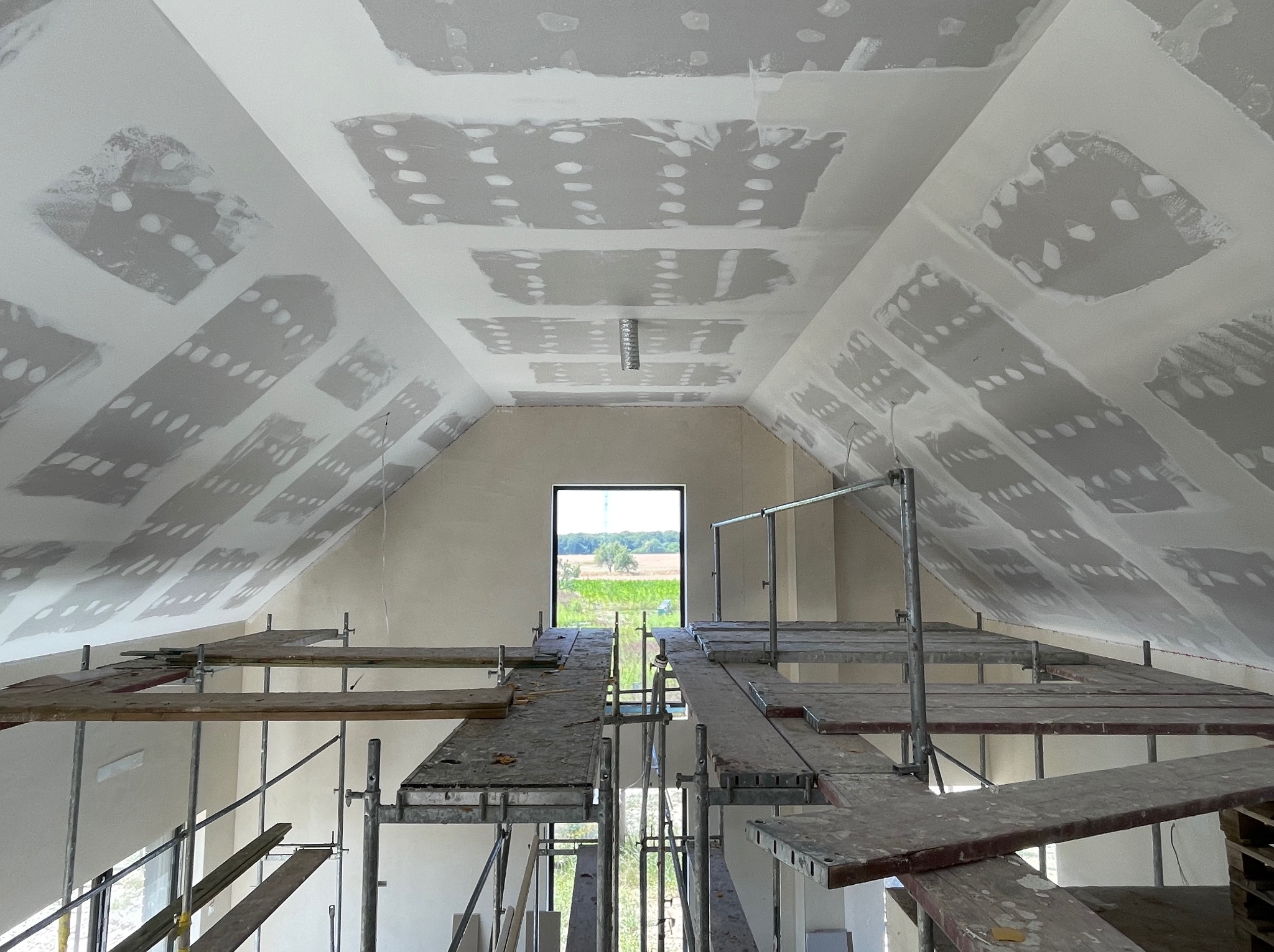 Poddasze w trakcie wykańczania: widoczne płyty karton-gips na skosach dachu, częściowo zaszpachlowane, rusztowanie w pomieszczeniu, okno z widokiem na zieleń.