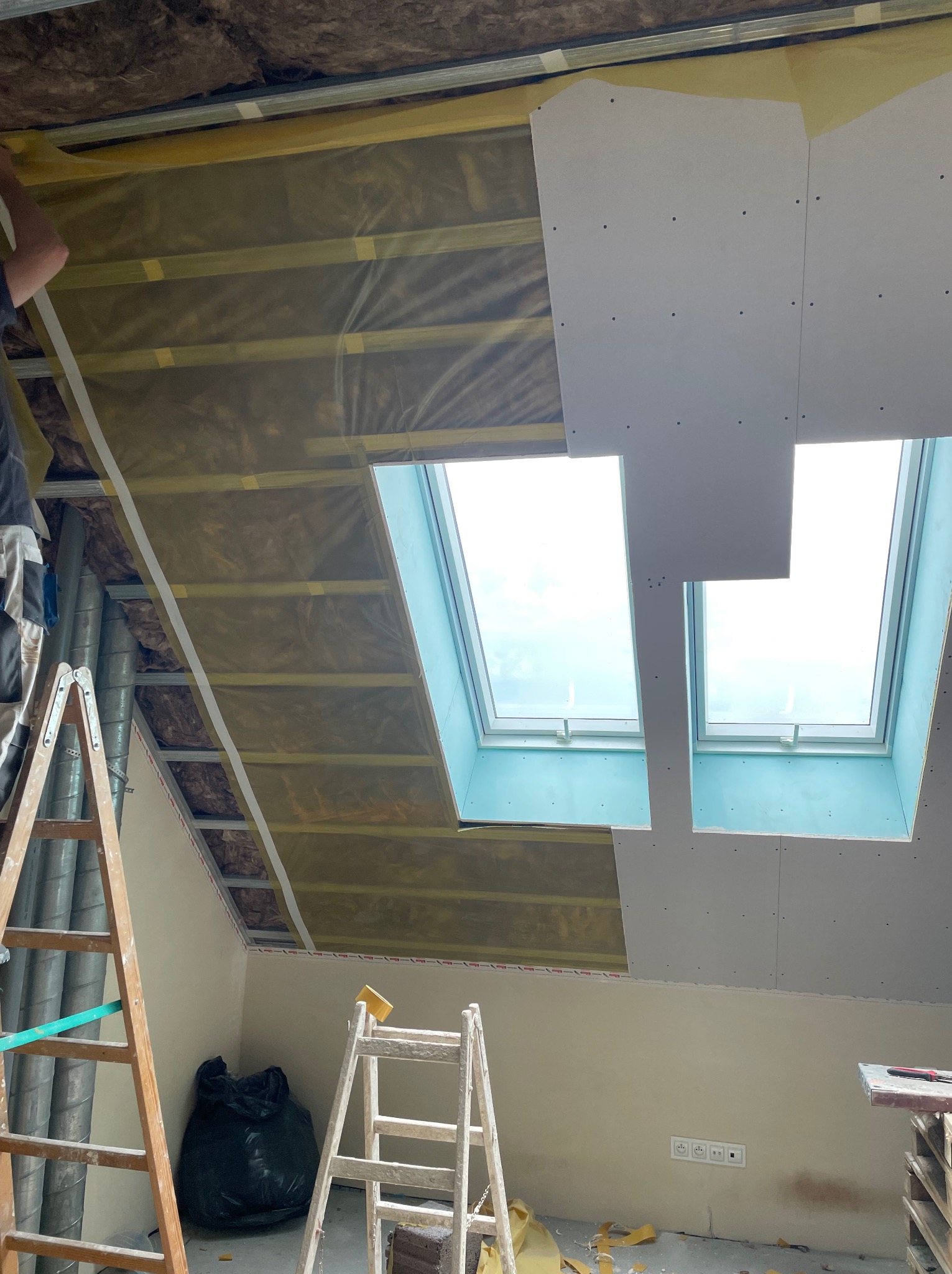 Montaż płyt G-K na poddaszu z oknami dachowymi w Rybiu. Widoczna izolacja i konstrukcja. Prace wykończeniowe w trakcie realizacji, drabiny i narzędzia w kadrze.