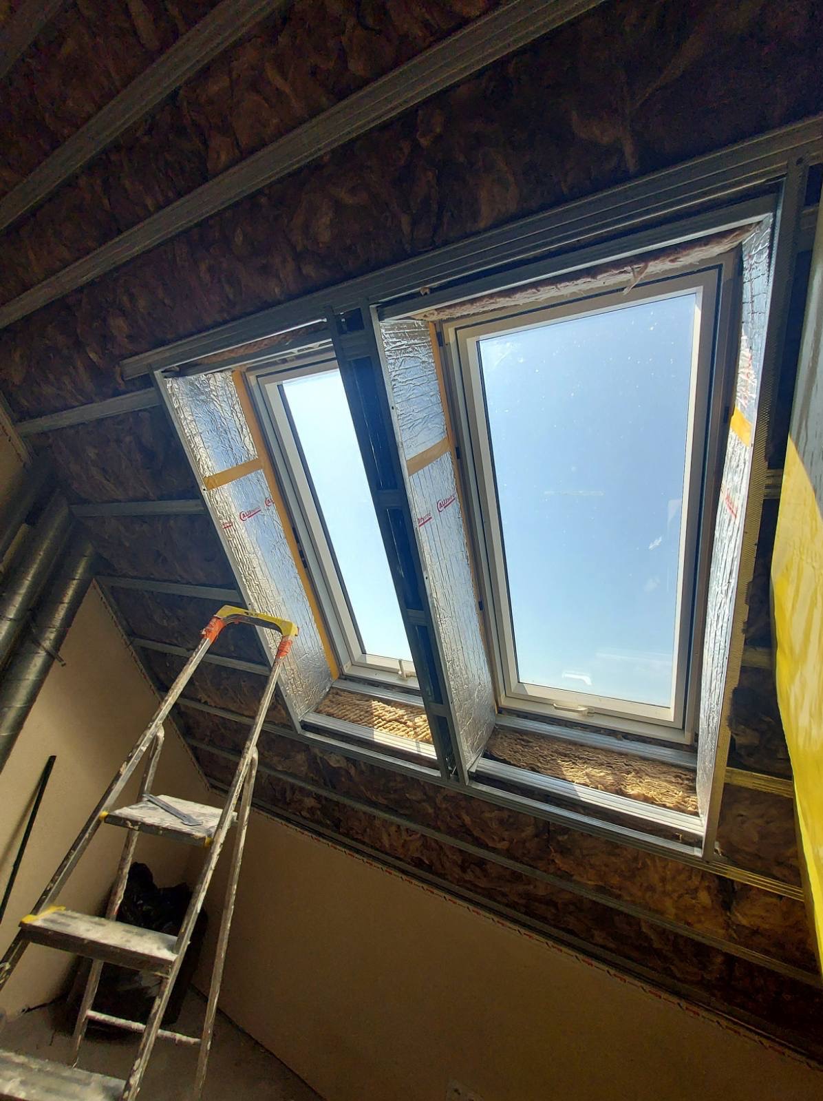 Dwa okna dachowe w trakcie montażu, widoczne ocieplenie wełną mineralną i metalowa konstrukcja. Ustawiona drabina malarska. Widok na błękitne niebo.