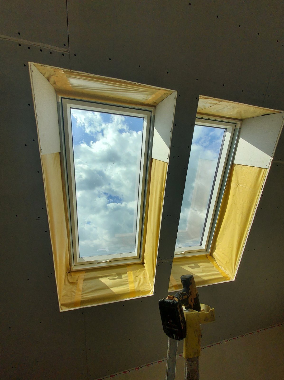 Montaż okien dachowych w płycie gipsowo-kartonowej, widoczne narzędzia na drabinie. Okna zabezpieczone folią, w tle błękitne niebo z chmurami.