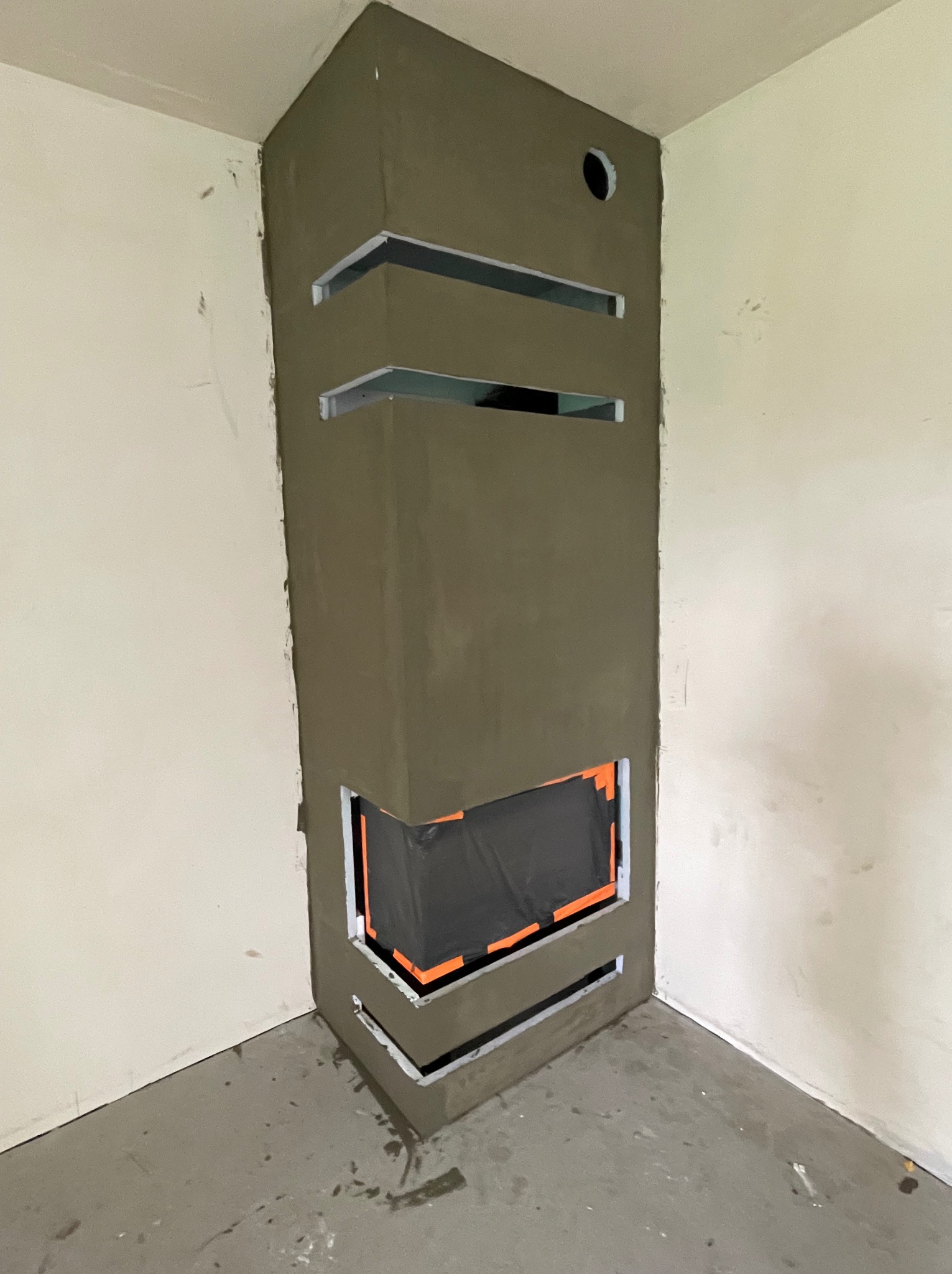 Wnętrze z obudową kominka w trakcie tynkowania, widoczne szczeliny wentylacyjne i otwór na rurę. Surowe ściany i podłoga w szarym odcieniu.