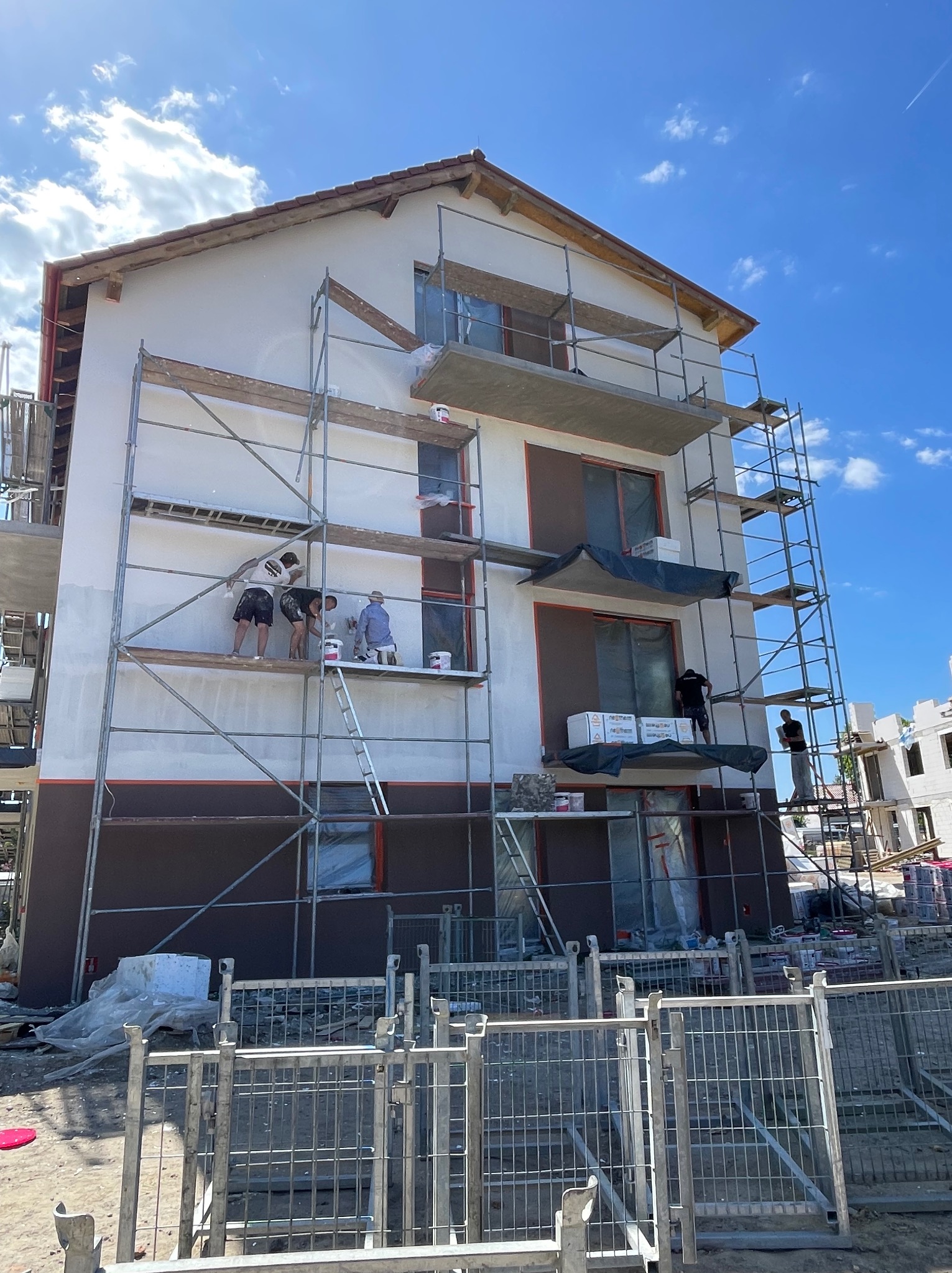 Ekipa maluje elewację dwukondygnacyjnego domu z rusztowania. Część ściany w kolorze białym, część brązowym. Błękitne niebo w tle. Widoczne narzędzia i materiały budowlane.