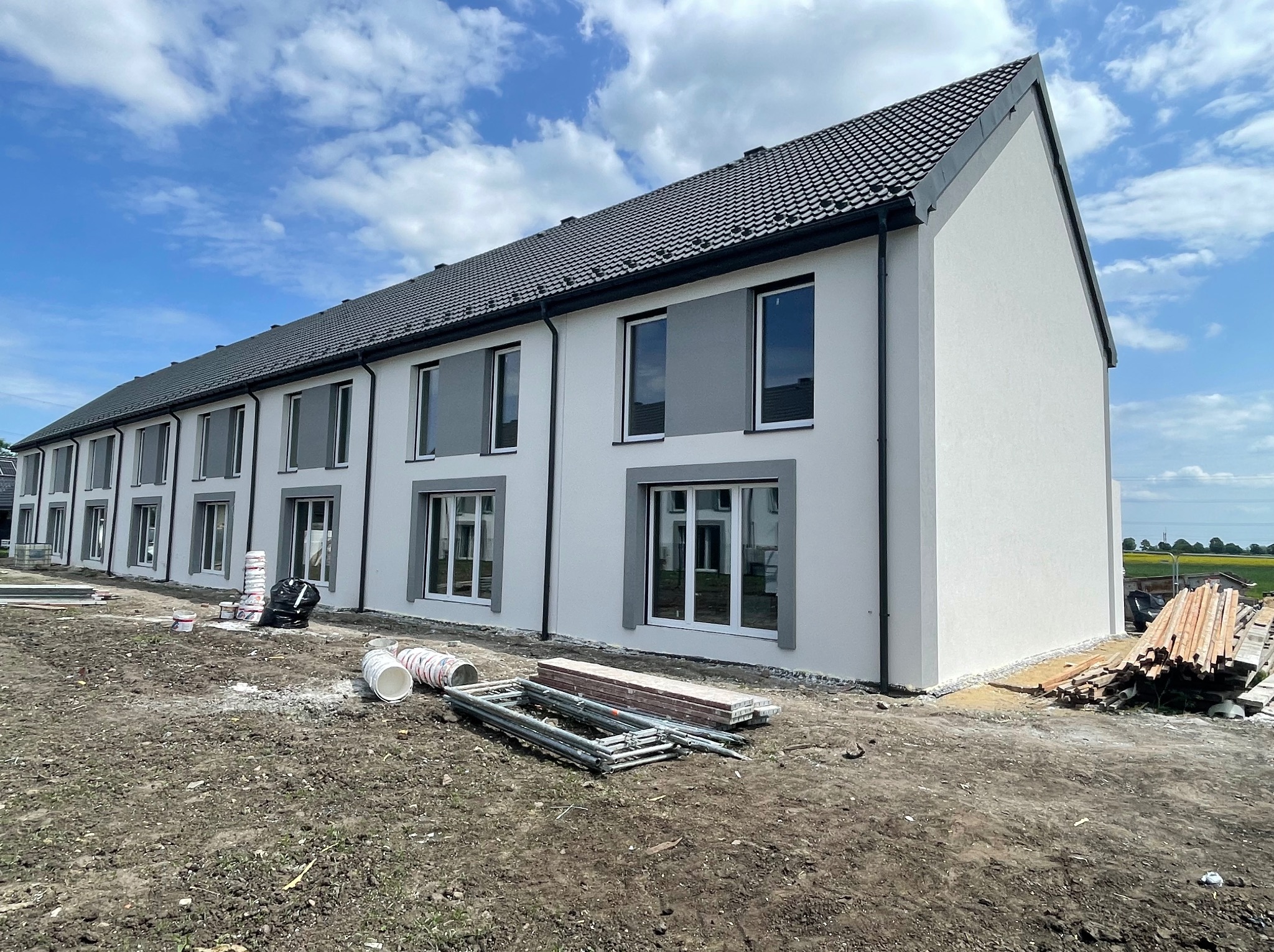 Nowoczesny szeregowiec po tynkowaniu elewacji, z widocznymi detalami wykończenia okien i dachu. Na pierwszym planie materiały budowlane i teren po pracach.