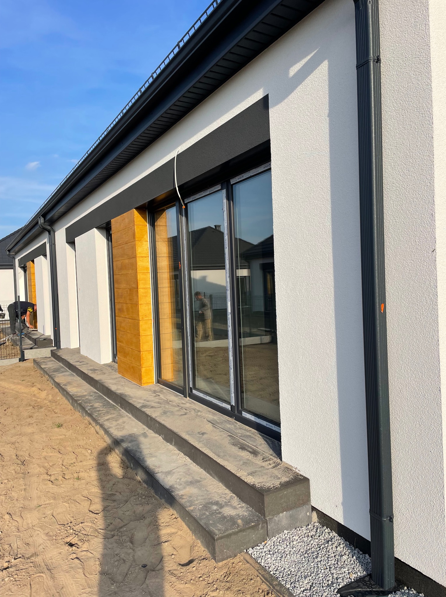 Elewacja domu jednorodzinnego z oknami przesuwnymi i drewnopodobnymi panelami. Widoczny fragment elewacji z fakturą baranka i czarną rynną spustową.