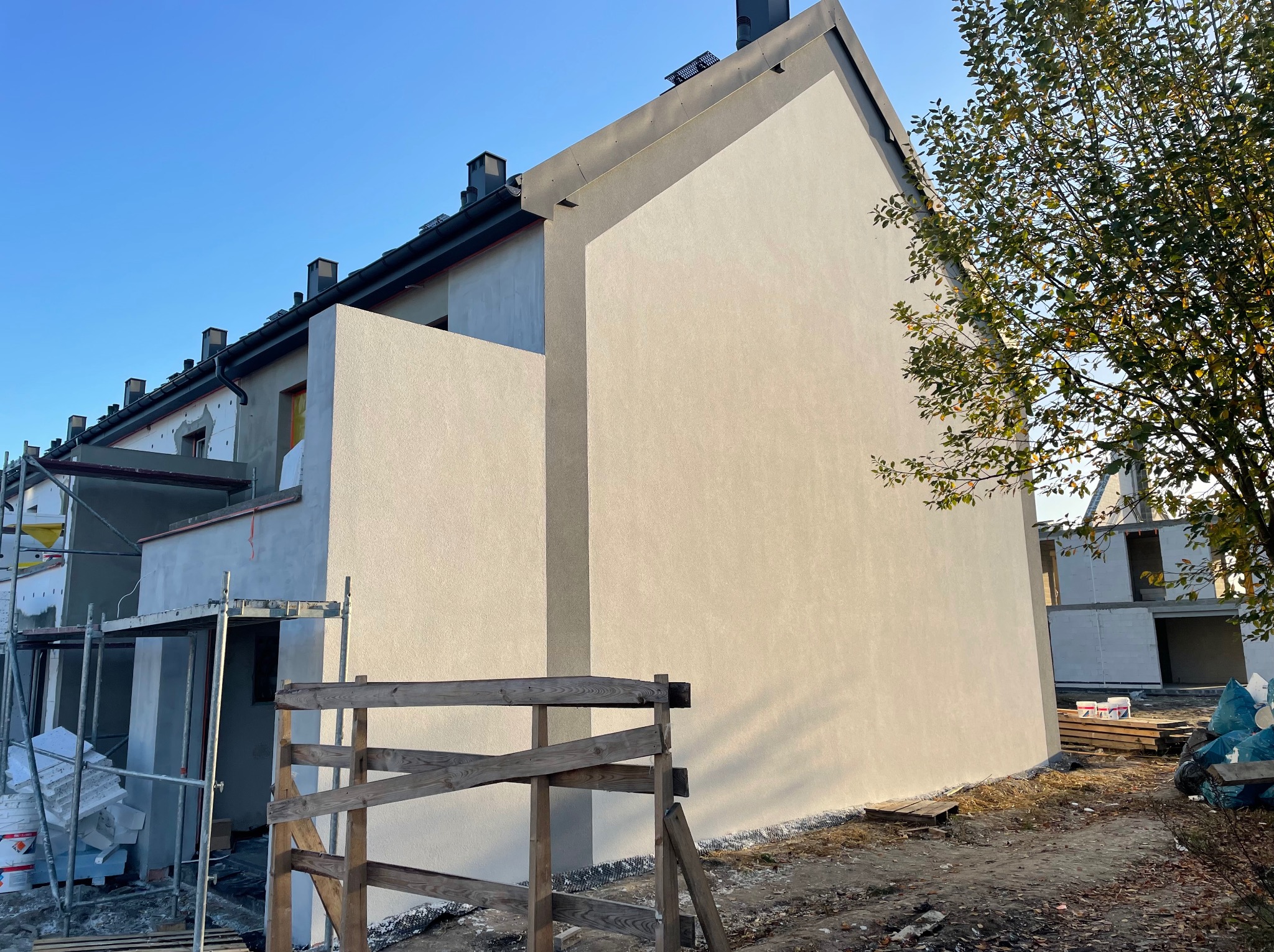 Nowo otynkowana elewacja domu jednorodzinnego w jasnym odcieniu, z widocznymi rusztowaniami i materiałami budowlanymi. Częściowo ocieplona ściana sąsiedniego budynku.