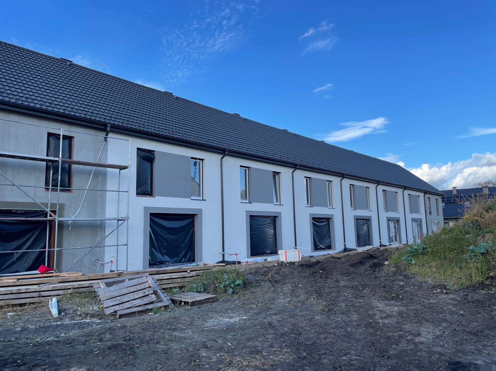 Szeregówka w trakcie prac elewacyjnych: szare ściany, czarne folie w oknach, rusztowania z boku. Na pierwszym planie ziemia i deski. Błękitne niebo w tle.