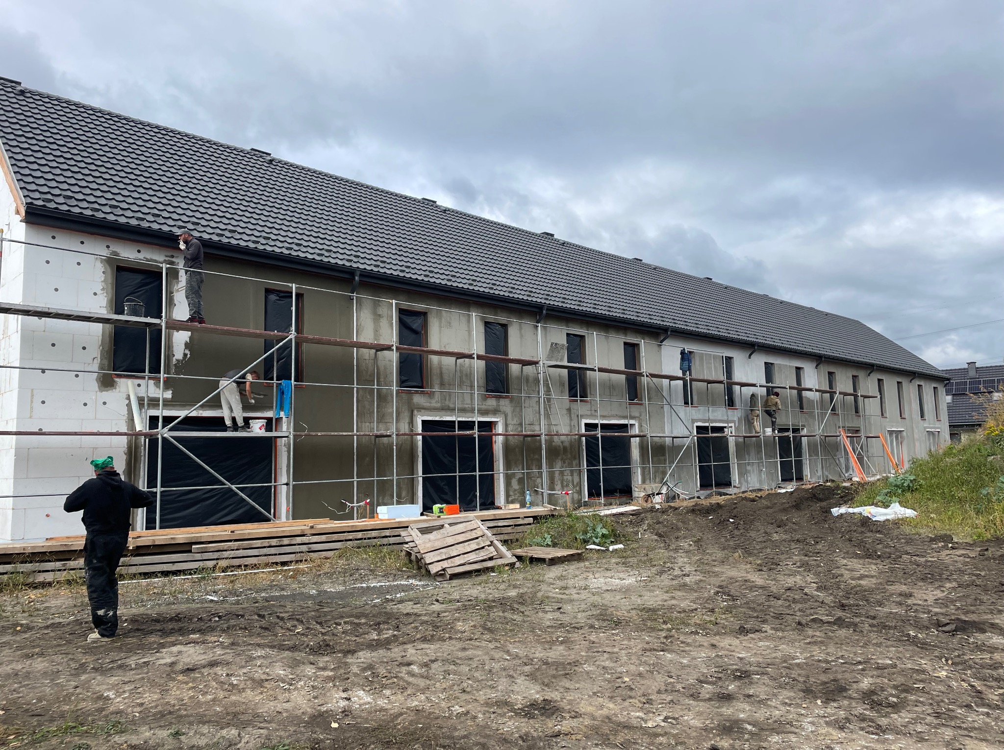 Długi budynek w trakcie ocieplania elewacji. Część ściany z białym styropianem, część z szarym tynkiem. Na rusztowaniach pracownicy. Pochmurne niebo.
