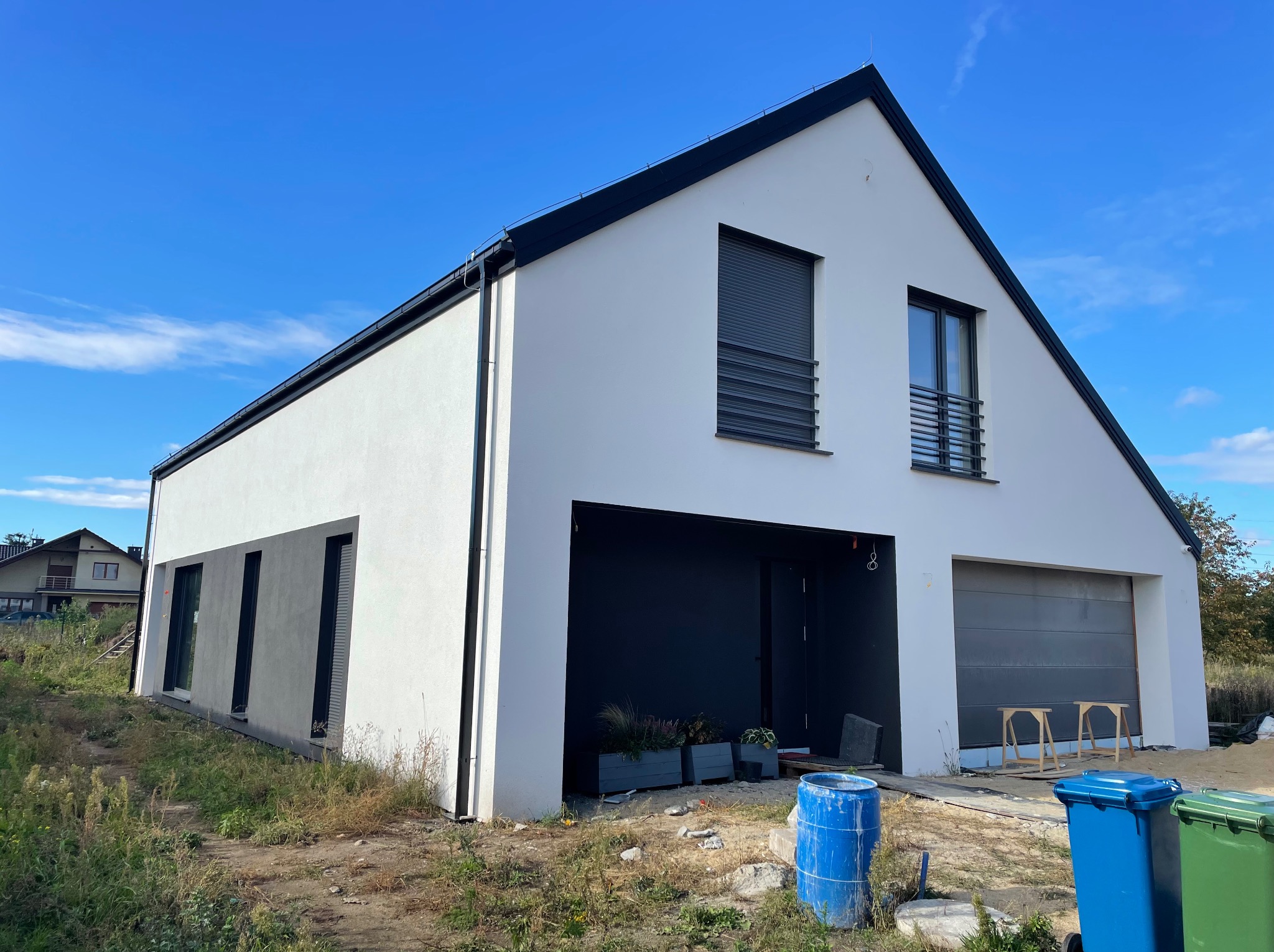 Nowoczesny dom jednorodzinny z białą elewacją i czarnymi akcentami, widok z zewnątrz w słoneczny dzień. Widoczne detale wykończenia i otoczenie budynku.