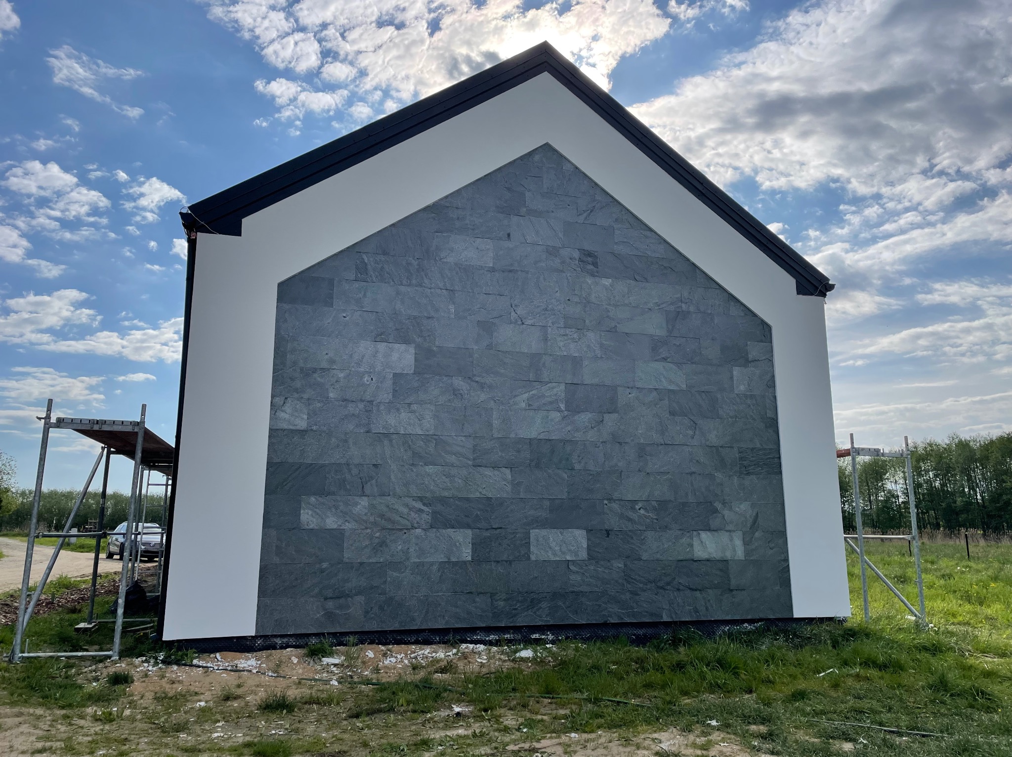 Elewacja domu z szarej, kamiennej okładziny, białe obramowanie, rusztowanie z boku. Słońce i chmury na niebie. Widok z dołu, podkreślający geometrię budynku.