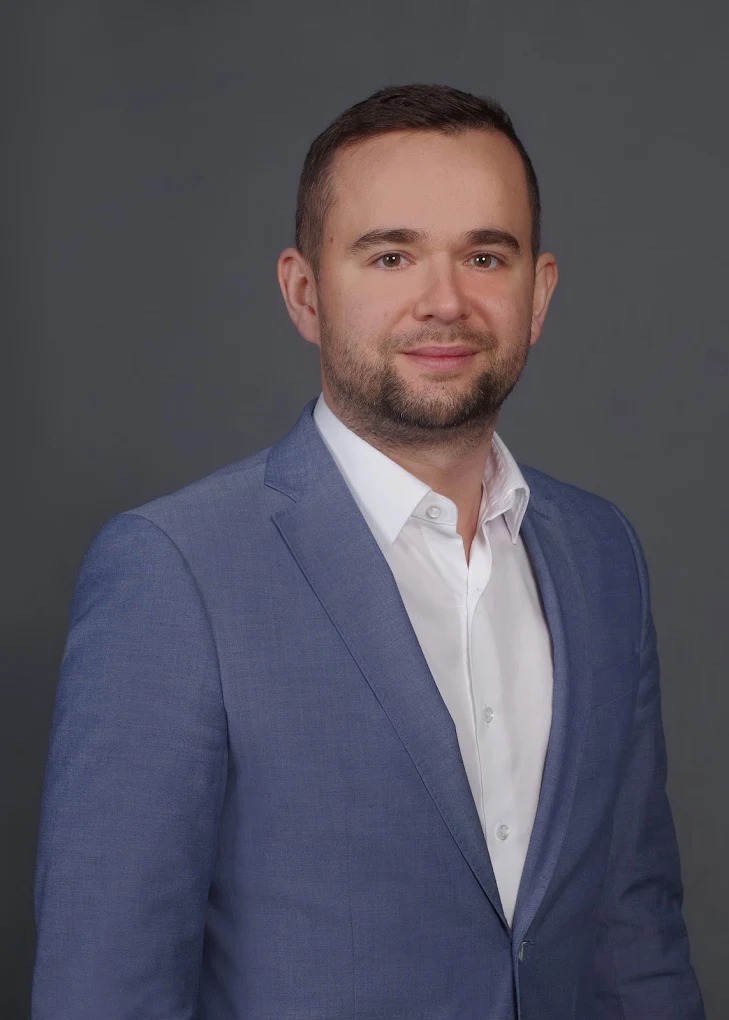 Portret mężczyzny w błękitnej marynarce i białej koszuli na szarym tle. Uśmiechnięty, z krótkimi włosami i lekkim zarostem. Profesjonalny wizerunek.