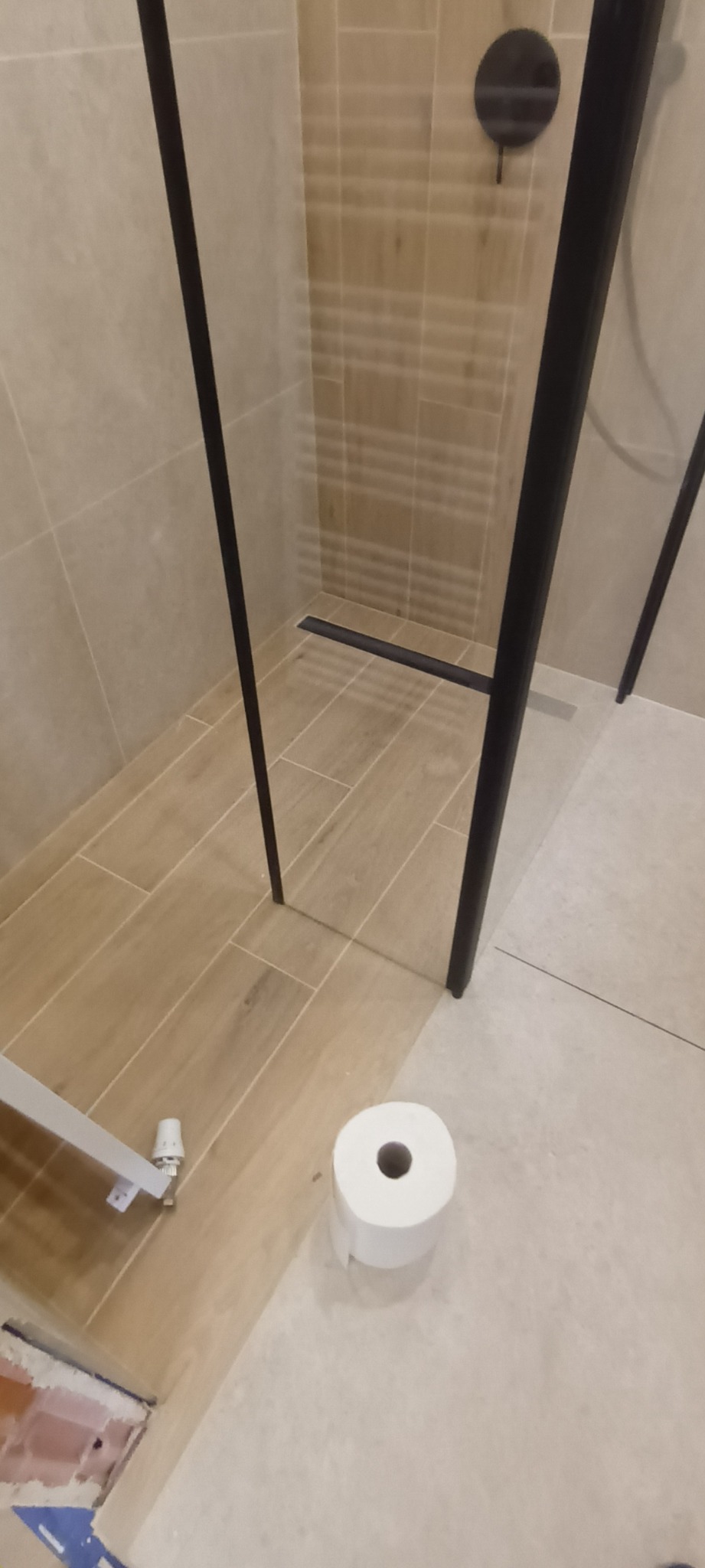 Beżowa łazienka z kabiną walk-in i odpływem liniowym. Widoczna rolka papieru toaletowego, płytki imitujące drewno i fragment niedokończonej obudowy wanny.