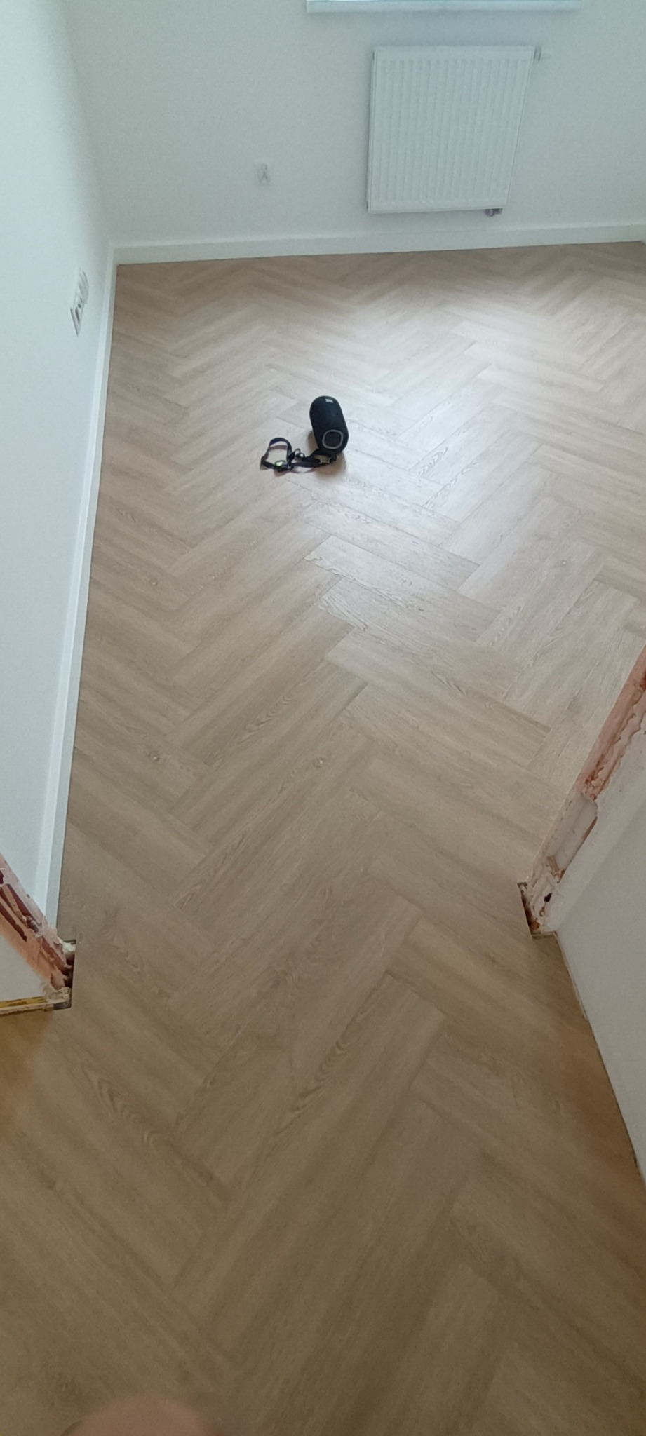 Jasna podłoga ułożona w jodełkę, widok z góry. Na podłodze leży głośnik i słuchawki. Ściany białe, widoczny grzejnik. Fragment futryny drzwi w prawym dolnym rogu.