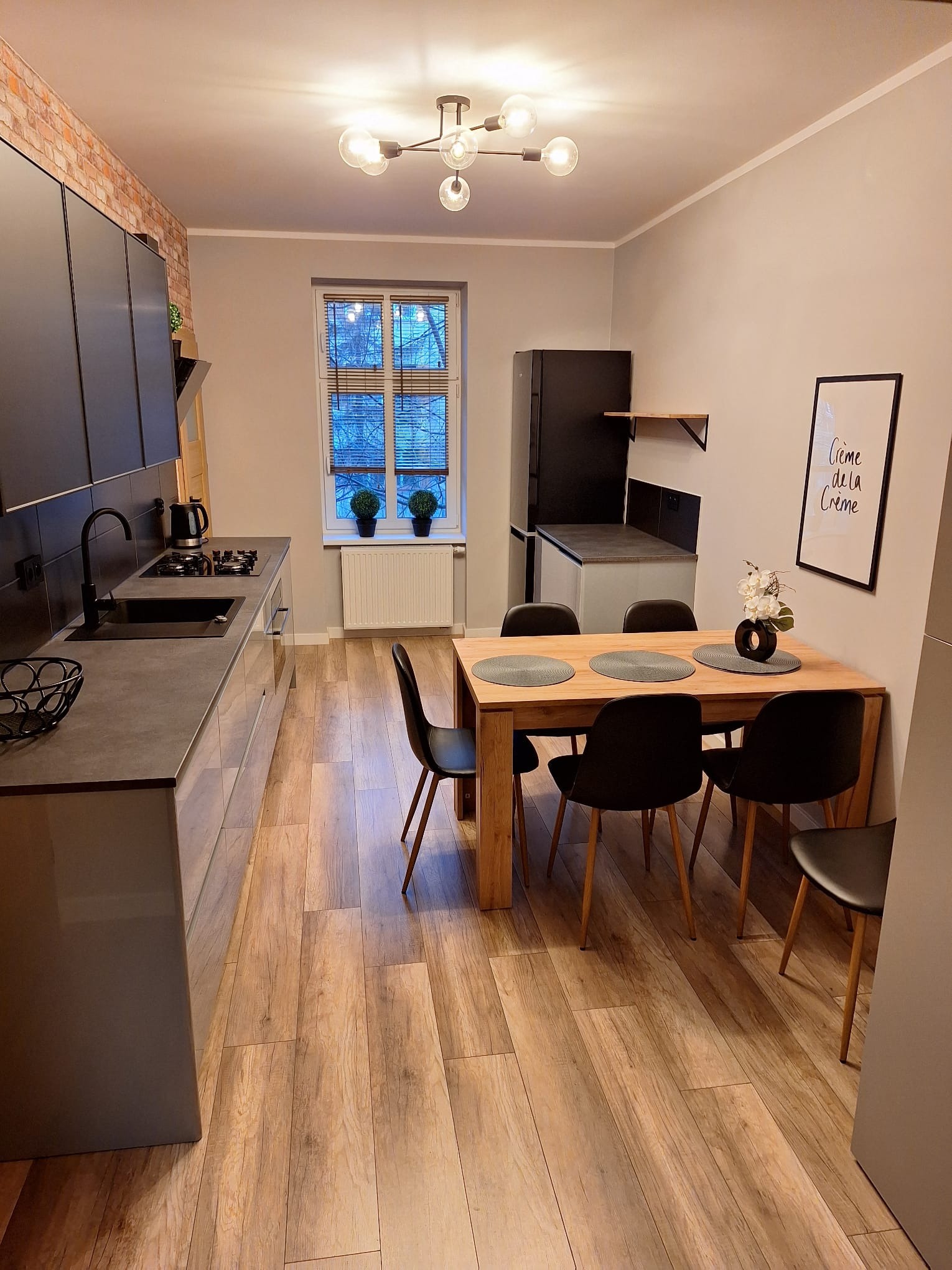 Nowoczesna kuchnia z jadalnią: szare szafki, drewniany stół z czarnymi krzesłami, parkiet imitujący drewno, ceglana ściana i designerska lampa sufitowa.