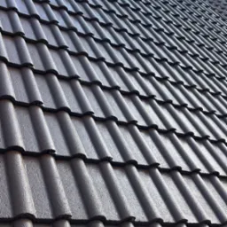 Ciemna, metalowa dachówka ceramiczna ułożona na dachu pod kątem, z widoczną fakturą i detalami łączeń. W tle fragment komina.