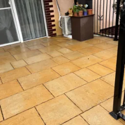 Żółta, prostokątna kostka brukowa na patio, widoczna barierka z czarnych metalowych prętów, drzwi balkonowe z zasłonami i donice z kwiatami.