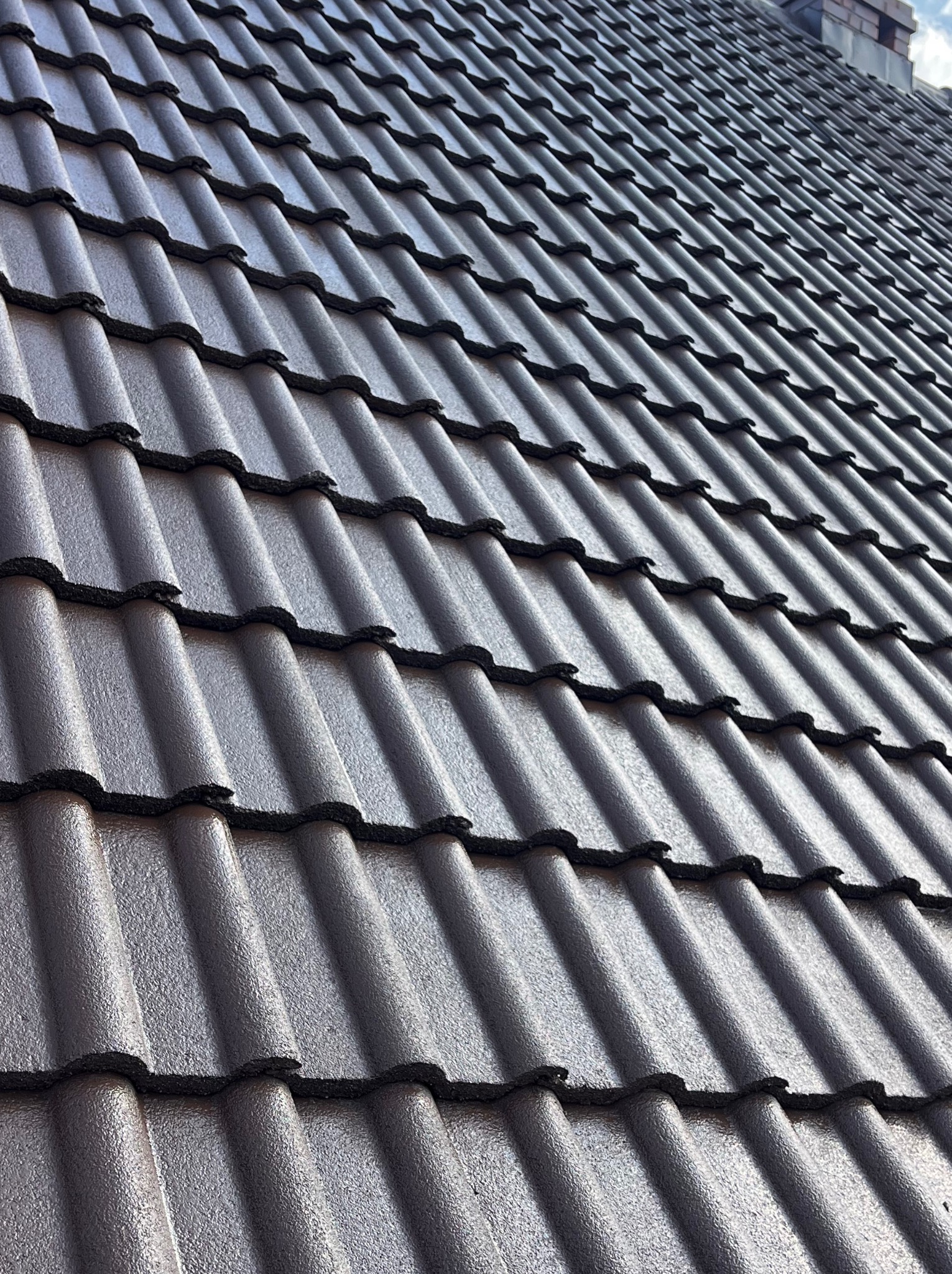 Ciemna, metalowa dachówka ceramiczna ułożona na dachu pod kątem, z widoczną fakturą i detalami łączeń. W tle fragment komina.