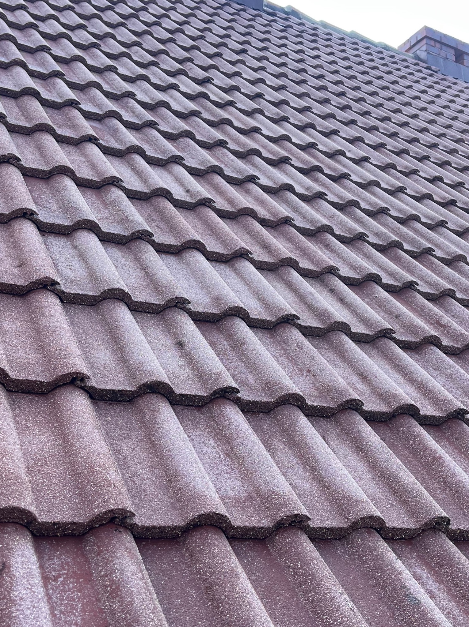 Czerwona dachówka bitumiczna ułożona na dachu, widok z dołu pod kątem. W tle komin. Dach jest lekko zabrudzony, co sugeruje potrzebę czyszczenia.