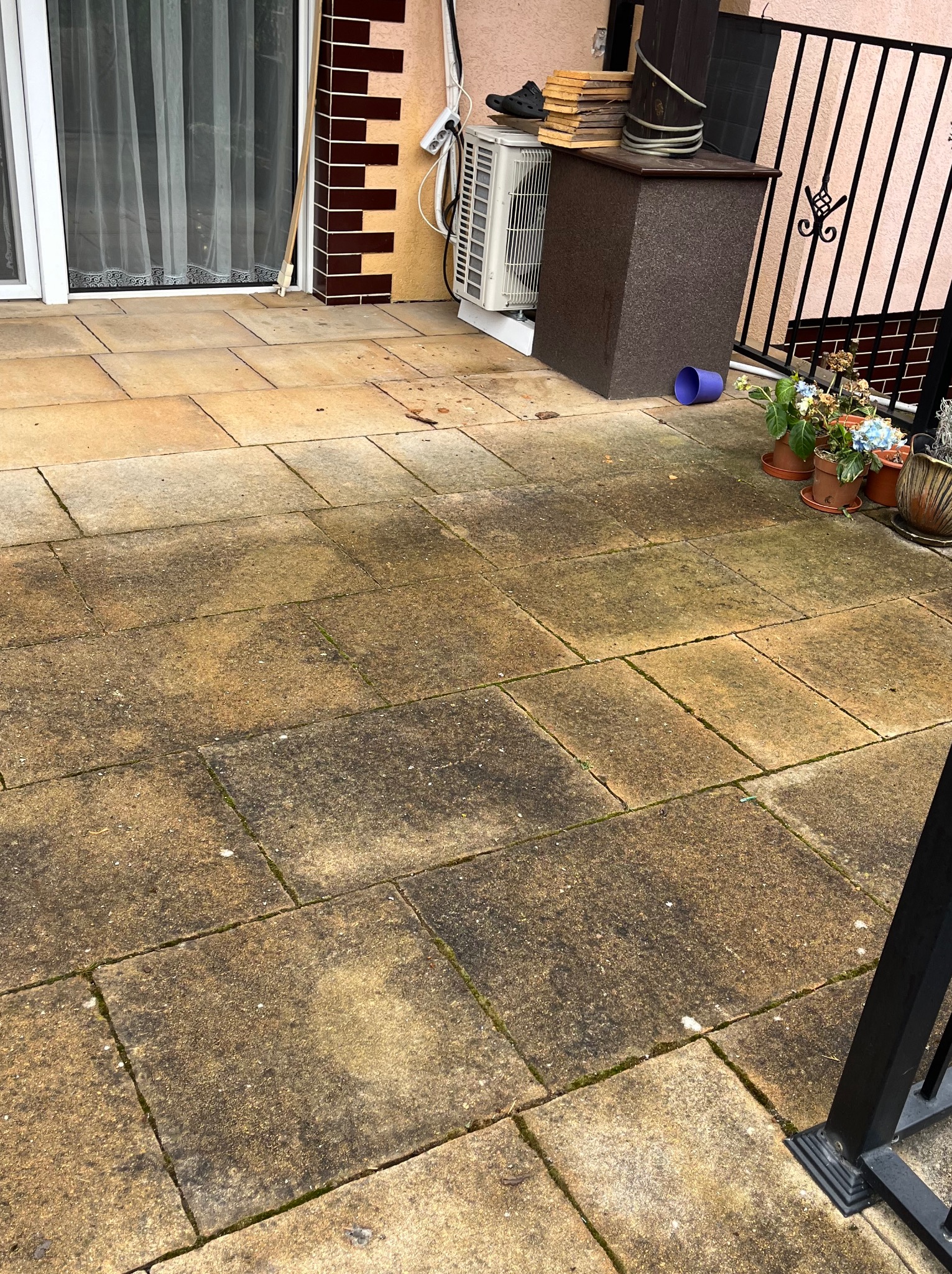 Brudna, kamienna posadzka tarasu w odcieniach beżu i brązu, wymagająca czyszczenia. W tle widoczne kwiaty w doniczkach, fragment balustrady i ściana budynku.