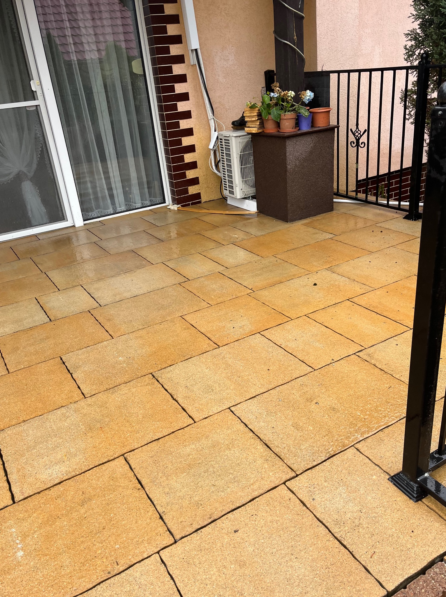 Żółta, prostokątna kostka brukowa na patio, widoczna barierka z czarnych metalowych prętów, drzwi balkonowe z zasłonami i donice z kwiatami.