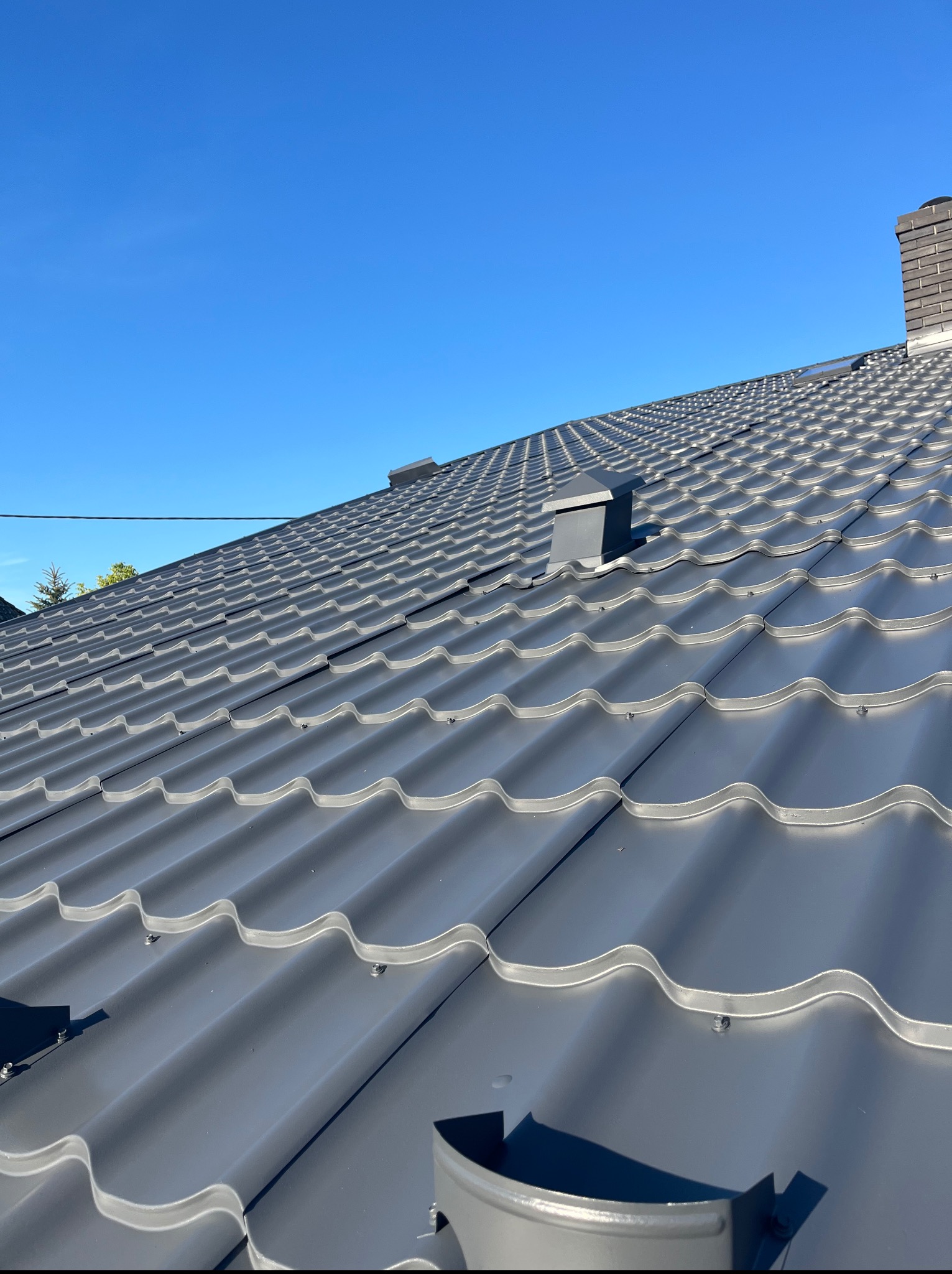 Szary dach z blachodachówki w słoneczny dzień, widok z góry, z kominem w tle. Widoczne mocowania blachy i fragment rynny.