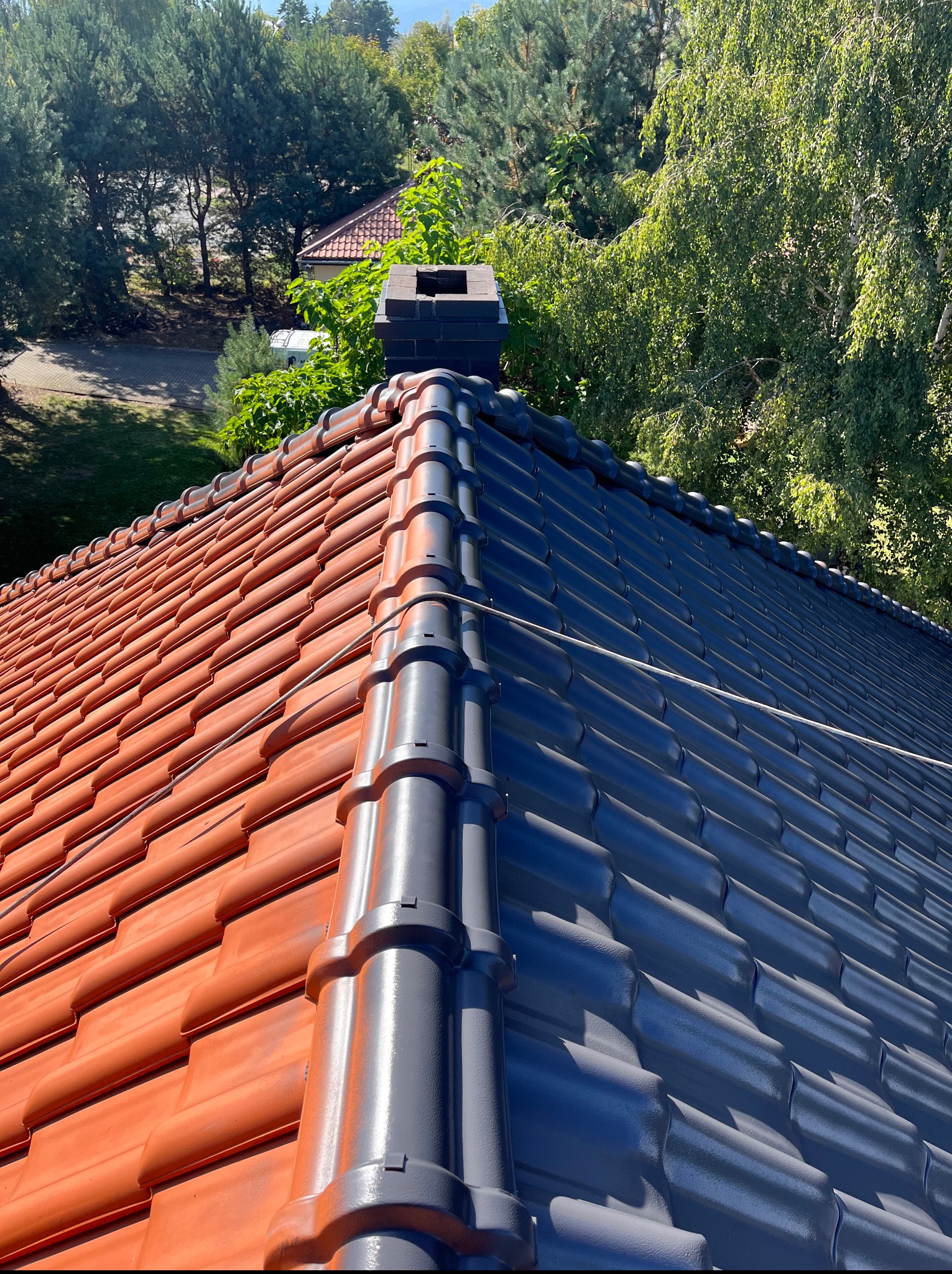 Częściowo pomalowany dach z ceglastą i szarą dachówką, widok z góry w Uciechowie. Połowa dachu odnowiona, komin w tle, drzewa i krzewy dookoła.