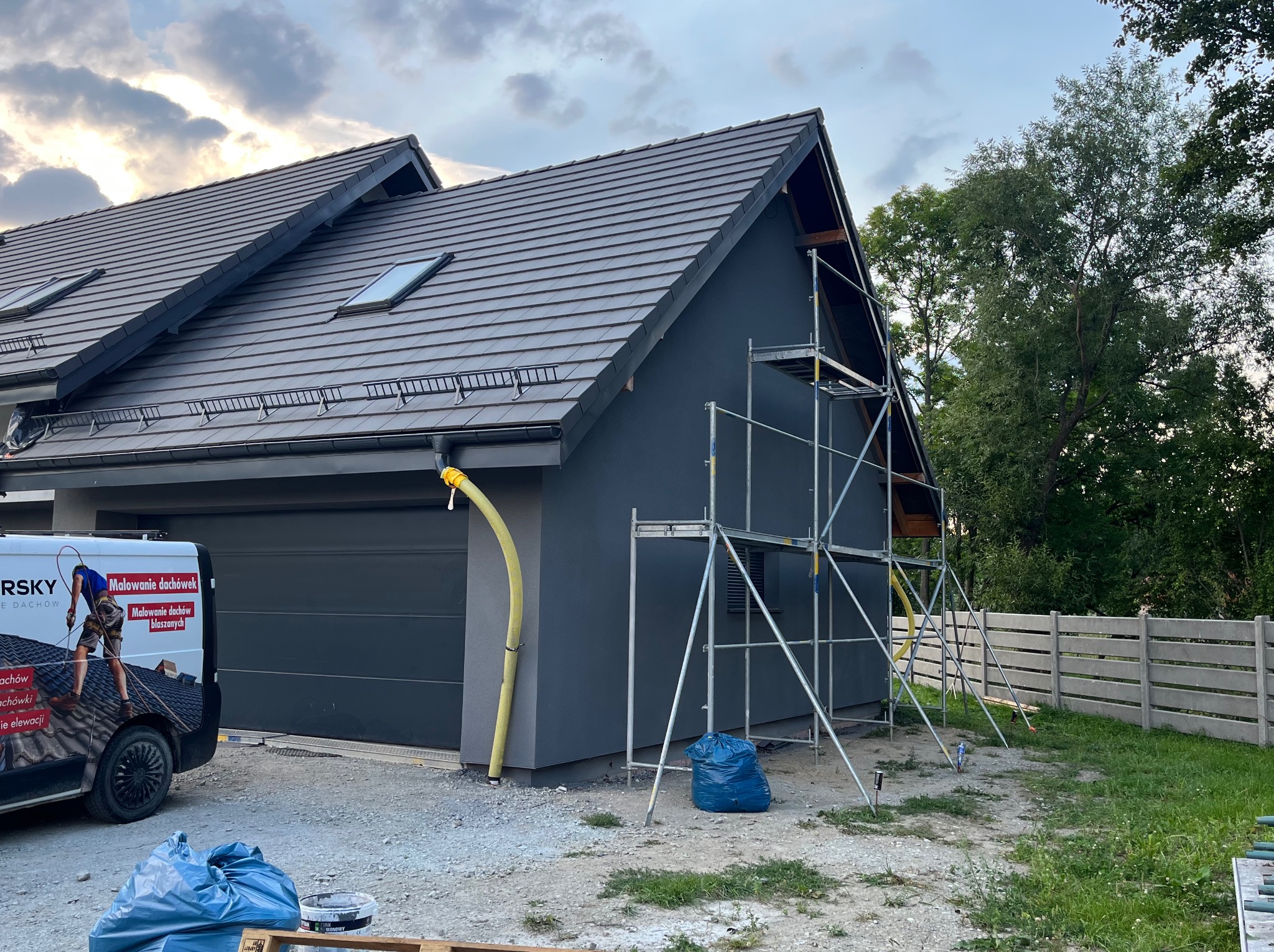 Dom z szarą elewacją i dachem, widoczne rusztowanie, worek na odpady i samochód firmowy z reklamą malowania dachów. Prace w toku.