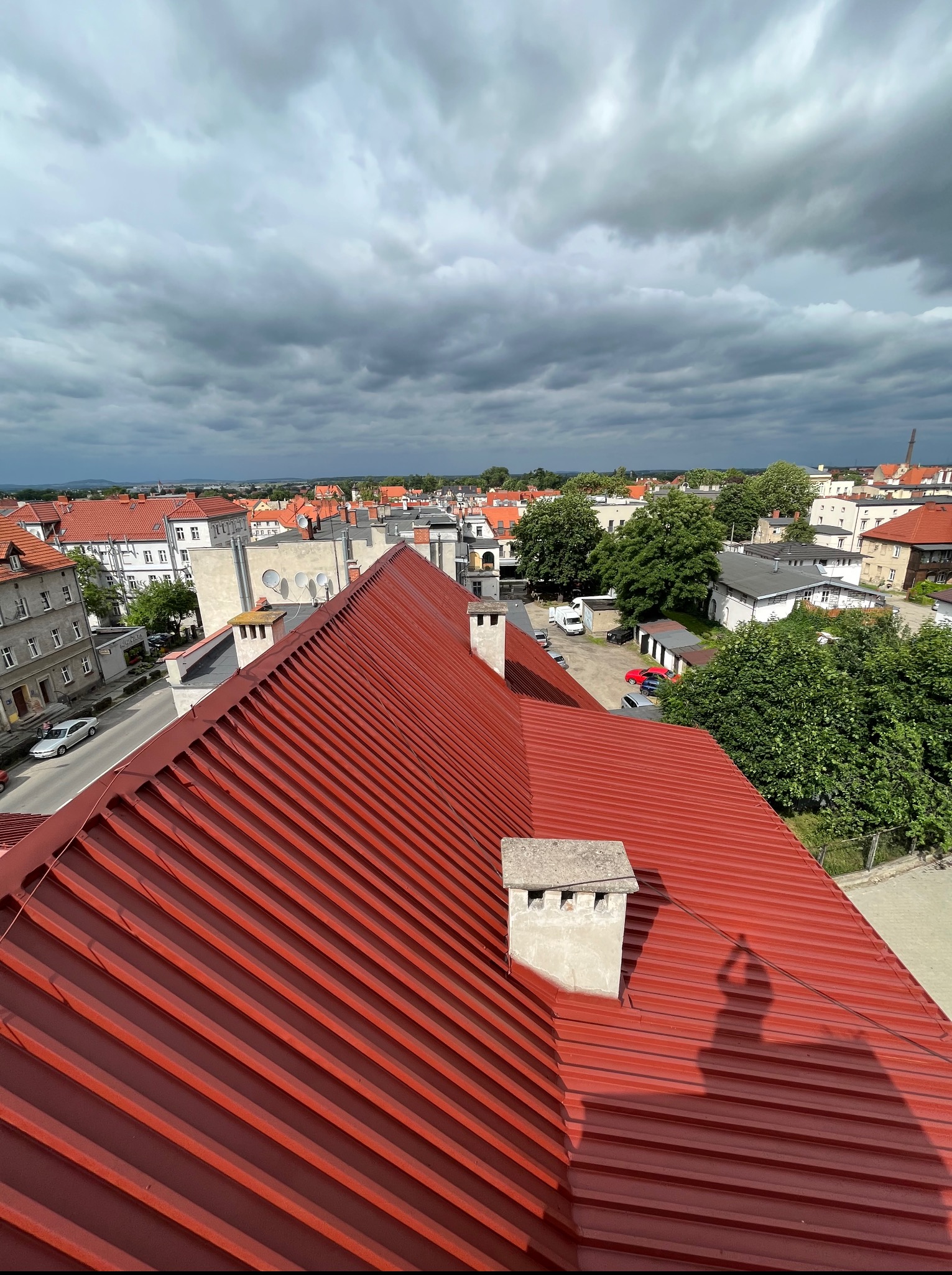 Czerwony, blaszany dach z kominami na tle miejskiego krajobrazu pod pochmurnym niebem. Widok z góry, ukazujący regularny wzór blachy i detale architektoniczne.