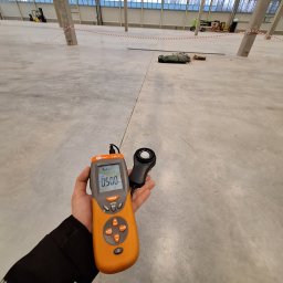 Damian Ławniczek-Wertepny - Pomiar natężenia oświetlenia luksomierzem na tle surowej posadzki w hali magazynowej z oknami i słupami. Na wyświetlaczu urządzenia widoczna wartość 0.500 lx.