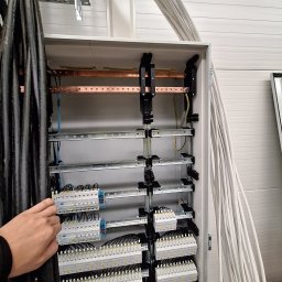 Damian Ławniczek-Wertepny - Rozdzielnia elektryczna w trakcie montażu: widoczne kable, szyny, zabezpieczenia. Ręka elektryka przytrzymuje przewody. Białe ściany w tle.
