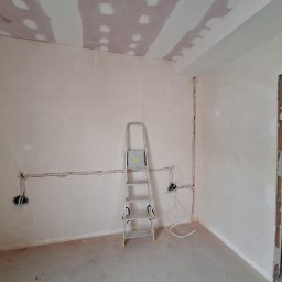 Damian Ławniczek-Wertepny - Wnętrze w trakcie remontu: ściany po gładzi, widoczne przewody elektryczne, drabina aluminiowa. Sufit z płyt gipsowych, częściowo zaszpachlowany. Otwór drzwiowy.