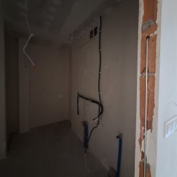 Damian Ławniczek-Wertepny - Wnętrze w trakcie remontu: widoczne przewody elektryczne, rury instalacji wodnej i fragment ściany z odsłoniętą cegłą. Surowy stan wykończenia, przygotowanie do dalszych prac.